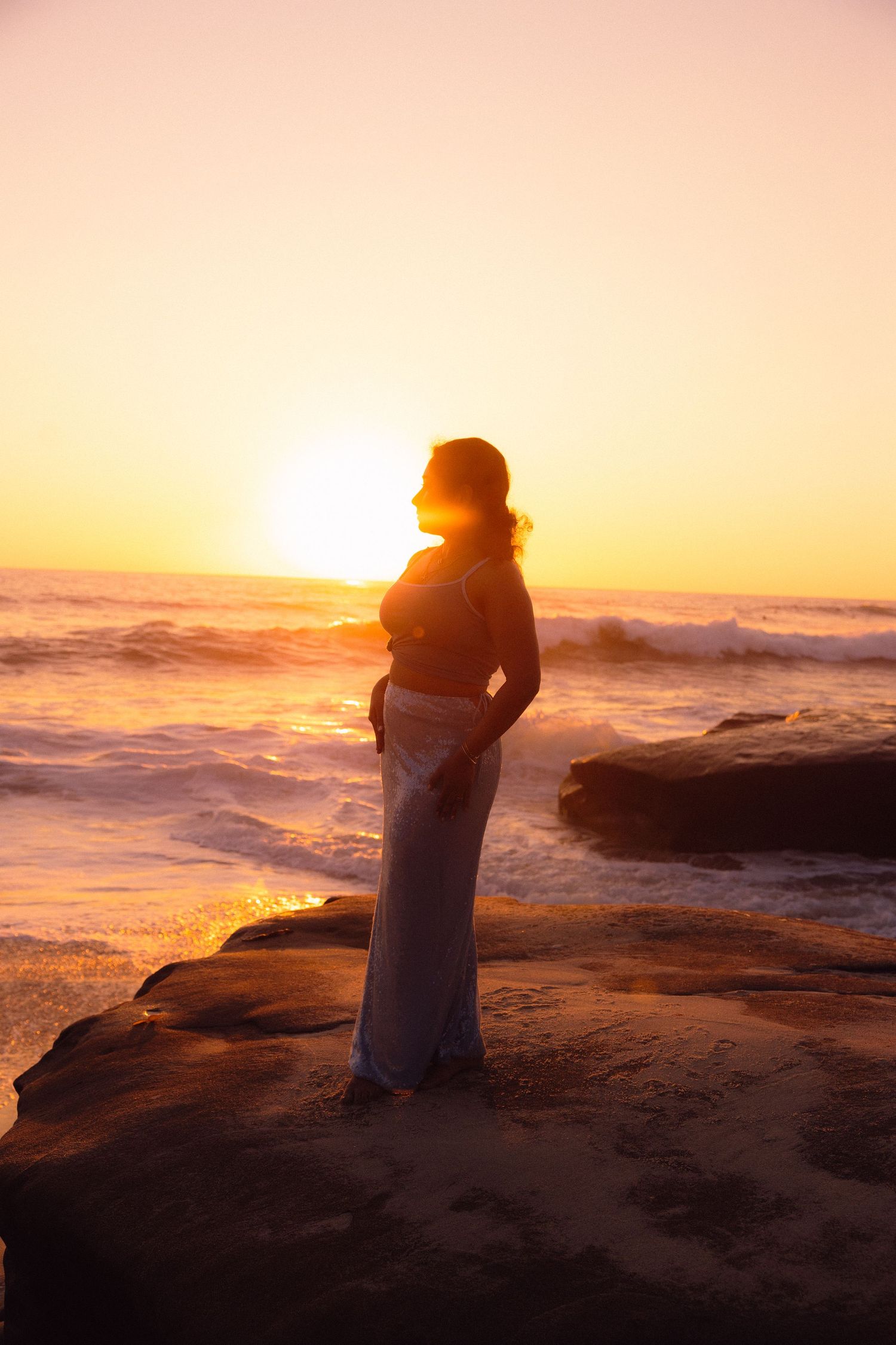 A silhouetted figure sits on rocky coastal cliffs during a vibrant orange sunset over crashing ocean waves.