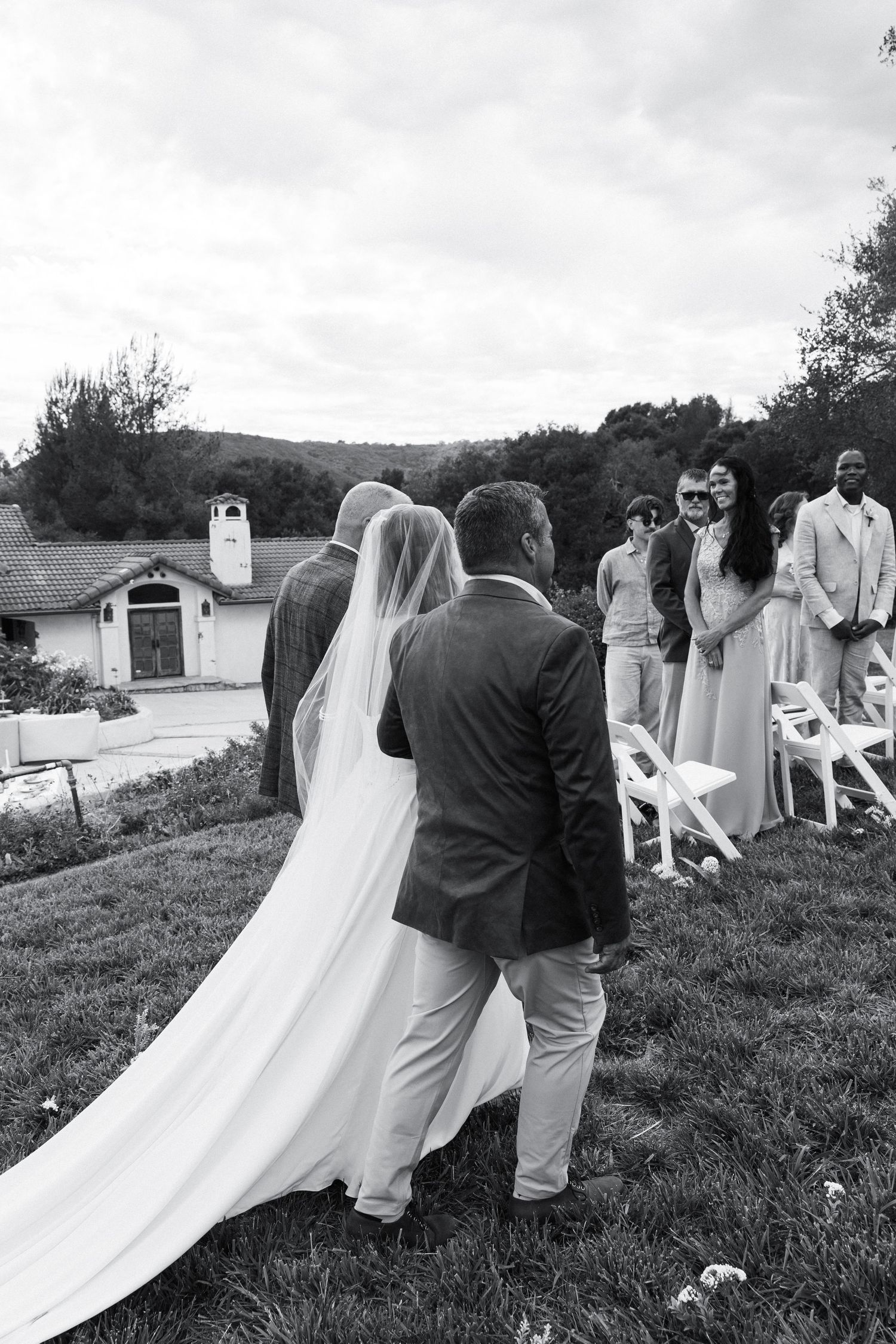 Black and white photo of a bride walking down the aisle with her escort at an outdoor wedding ceremony.