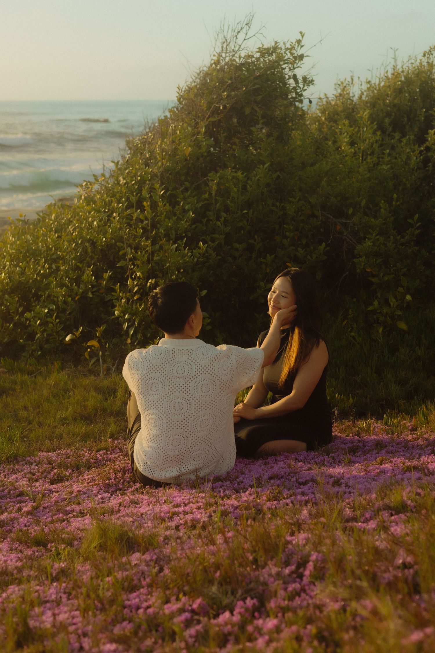Two people sit together in a field of purple wildflowers at sunset with bushes in the background.