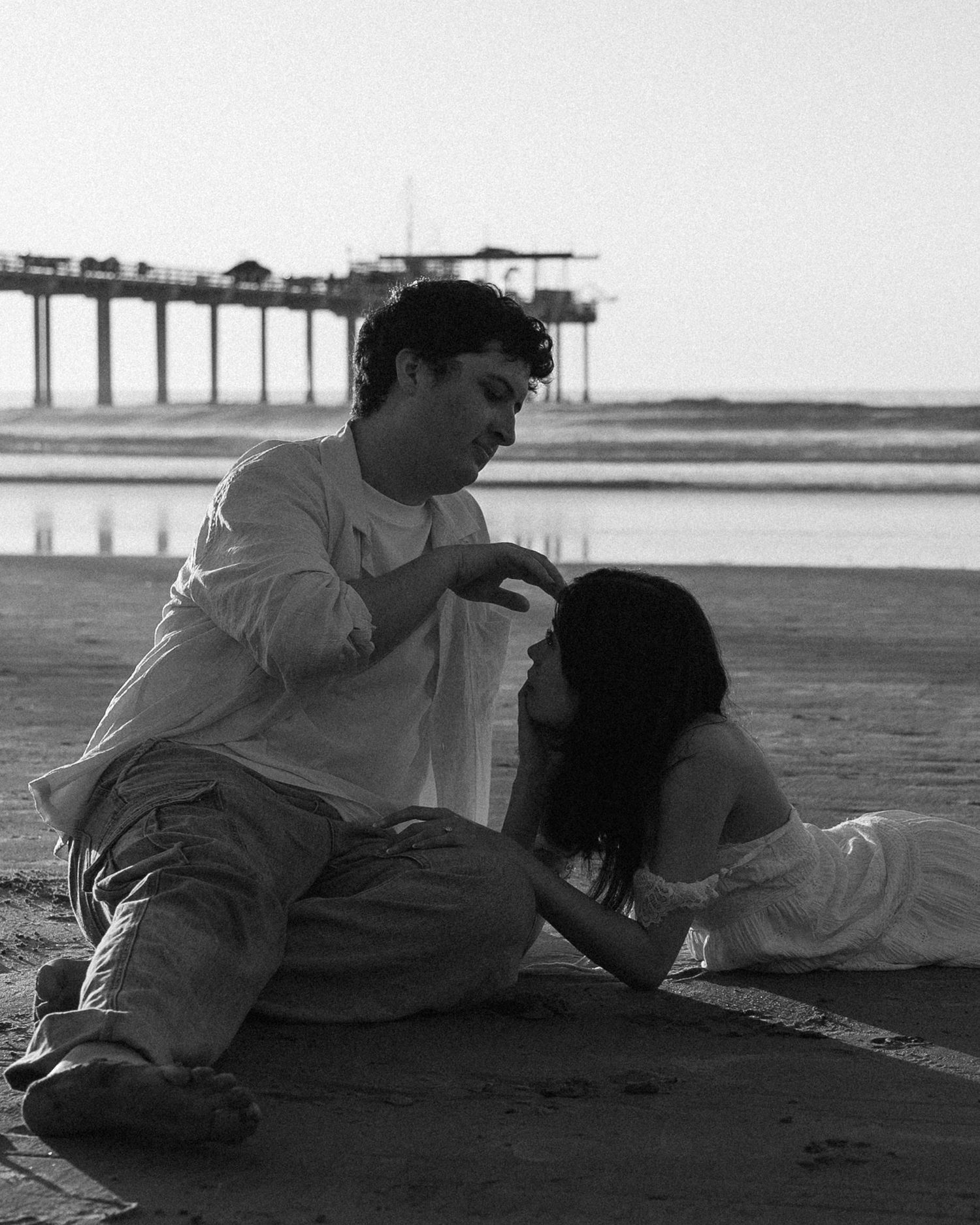 Two figures resting on a sandy beach with a pier visible in the background.