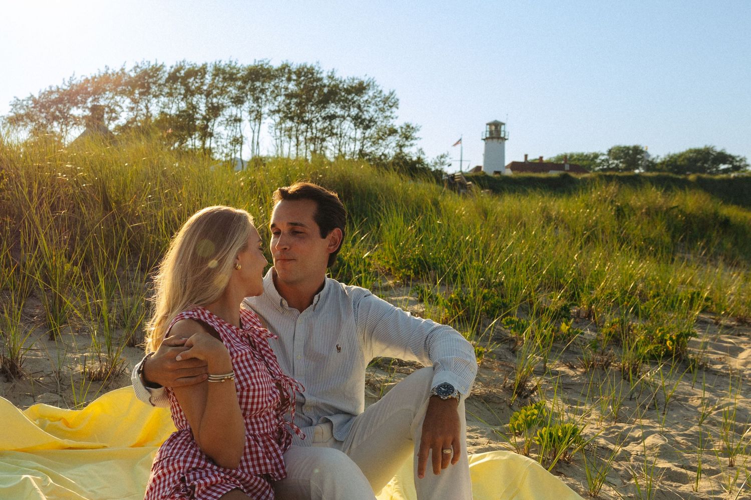 Couple sitting on sandy beach dunes near a lighthouse, surrounded by tall grass in warm golden sunlight.