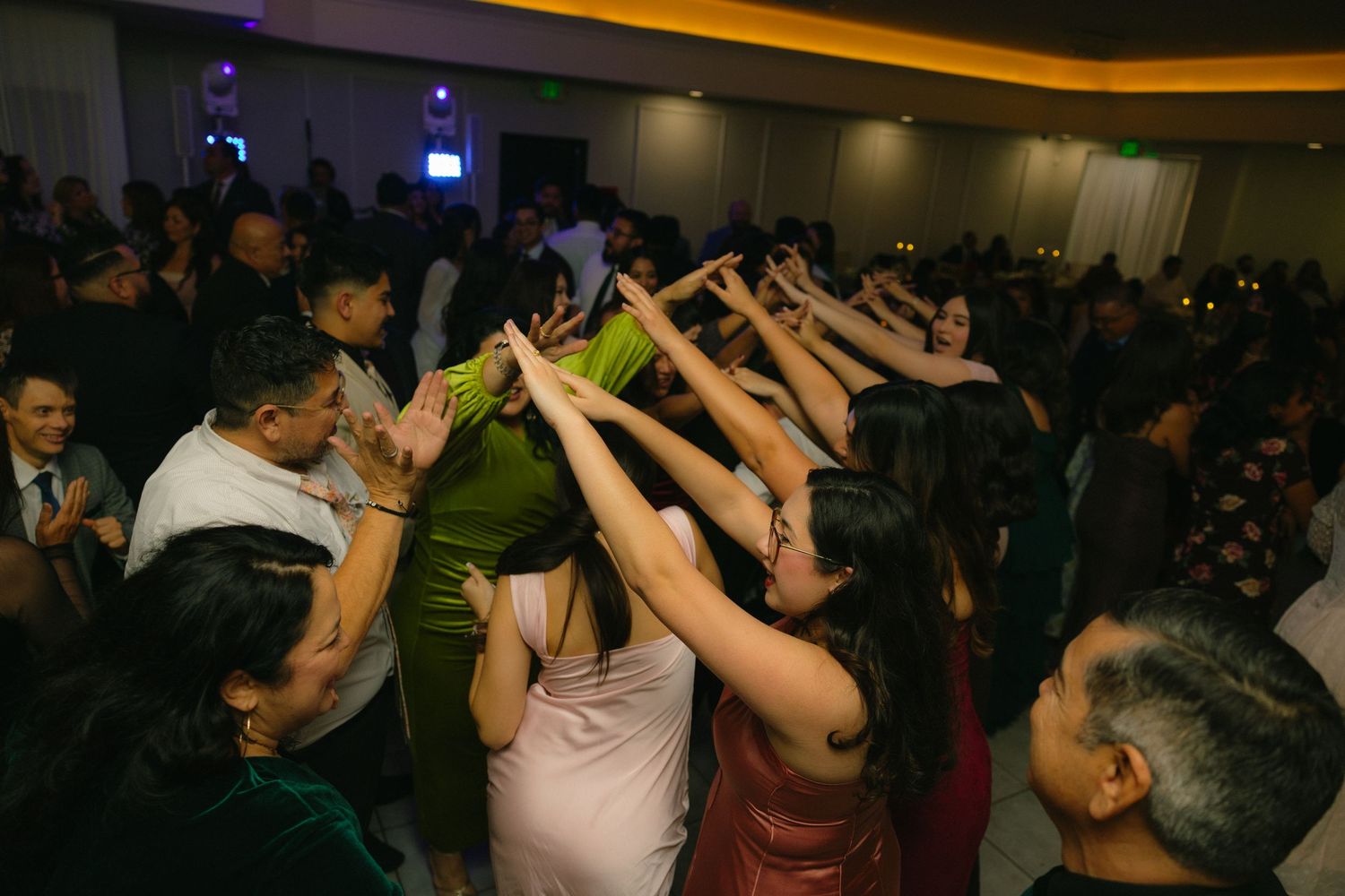 Wedding guests dancing and celebrating with raised arms on a crowded dance floor under ambient lighting.