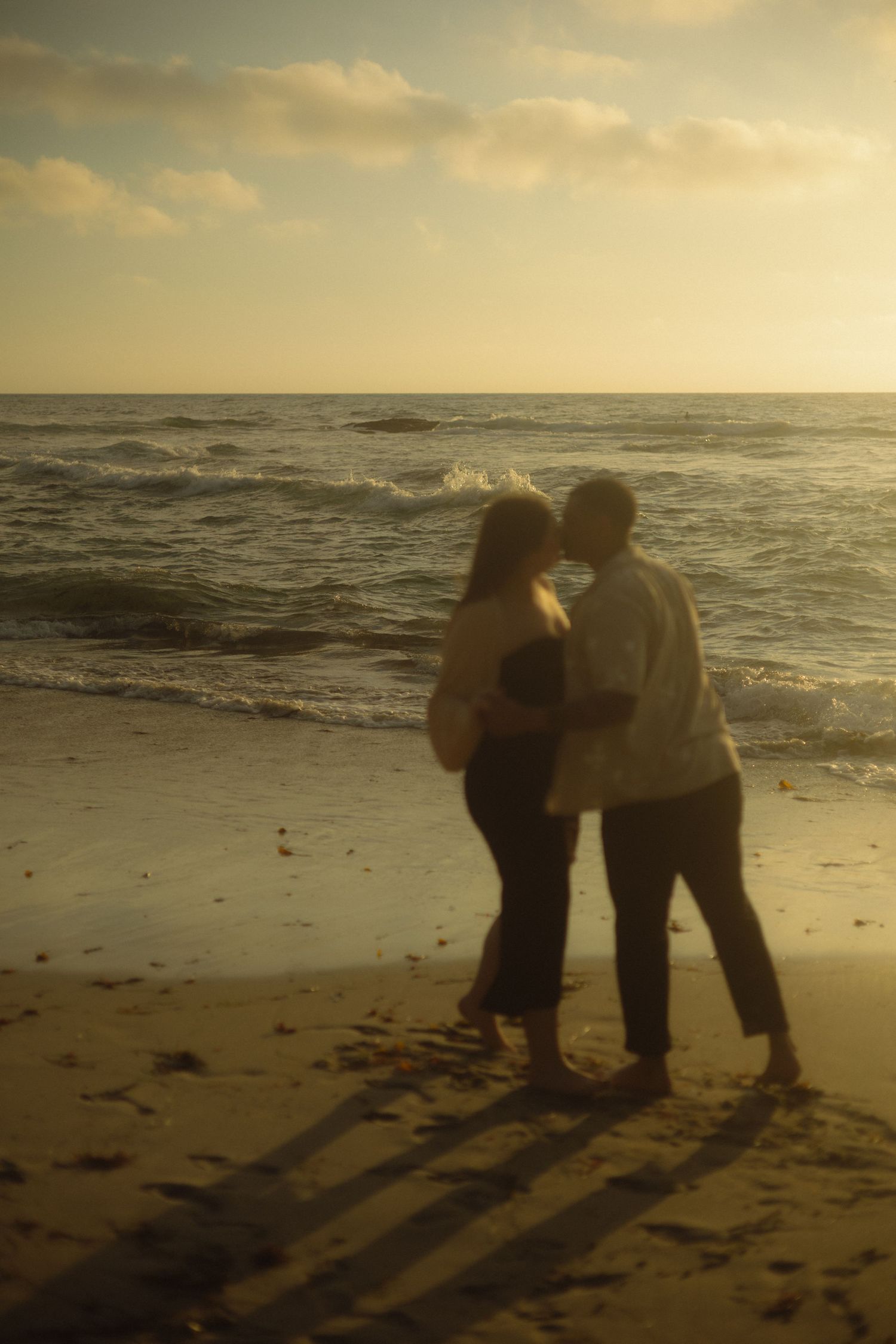 Two silhouettes embrace while watching the sunset over ocean waves on a serene beach.