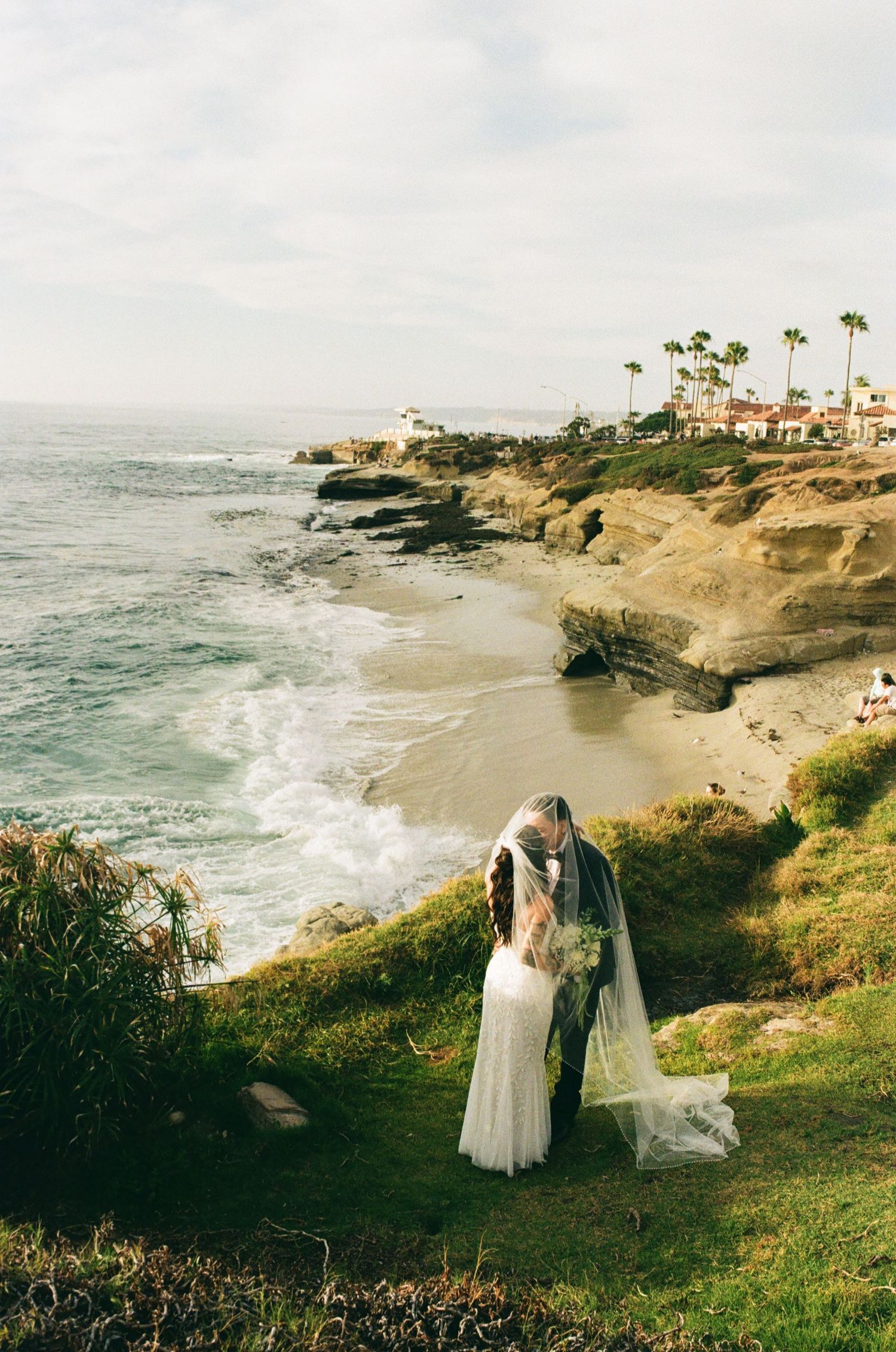 A bride and groom share a romantic kiss on a coastal cliff overlooking a sandy beach and ocean in La Jolla, California.