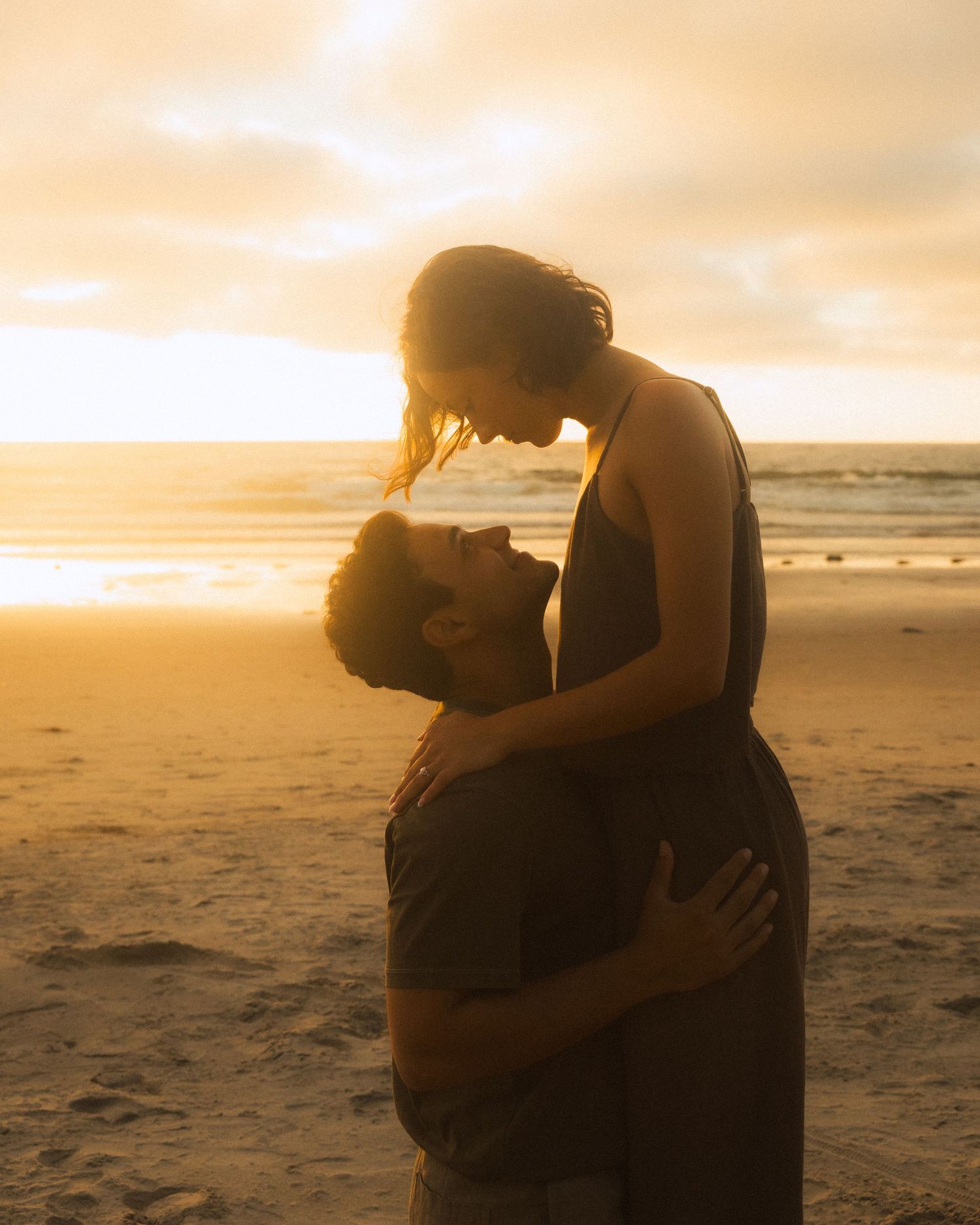Two silhouettes share an intimate embrace on a beach at sunset with golden light illuminating the scene.