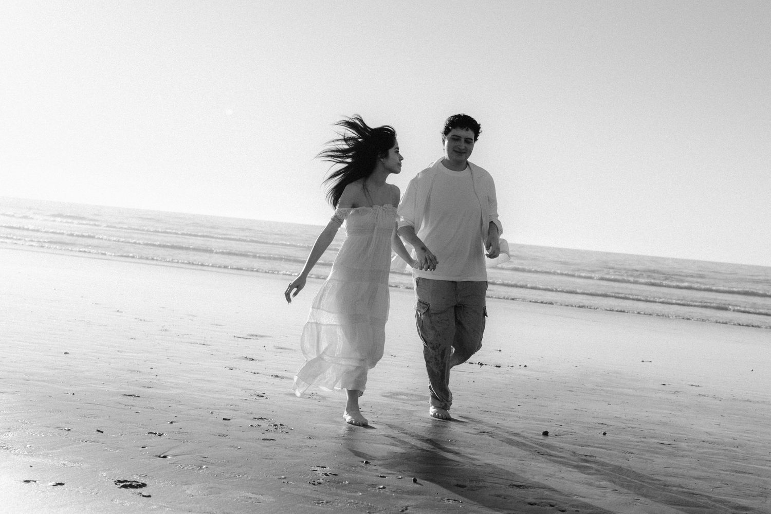 A couple runs together along a misty beach shoreline in black and white.
