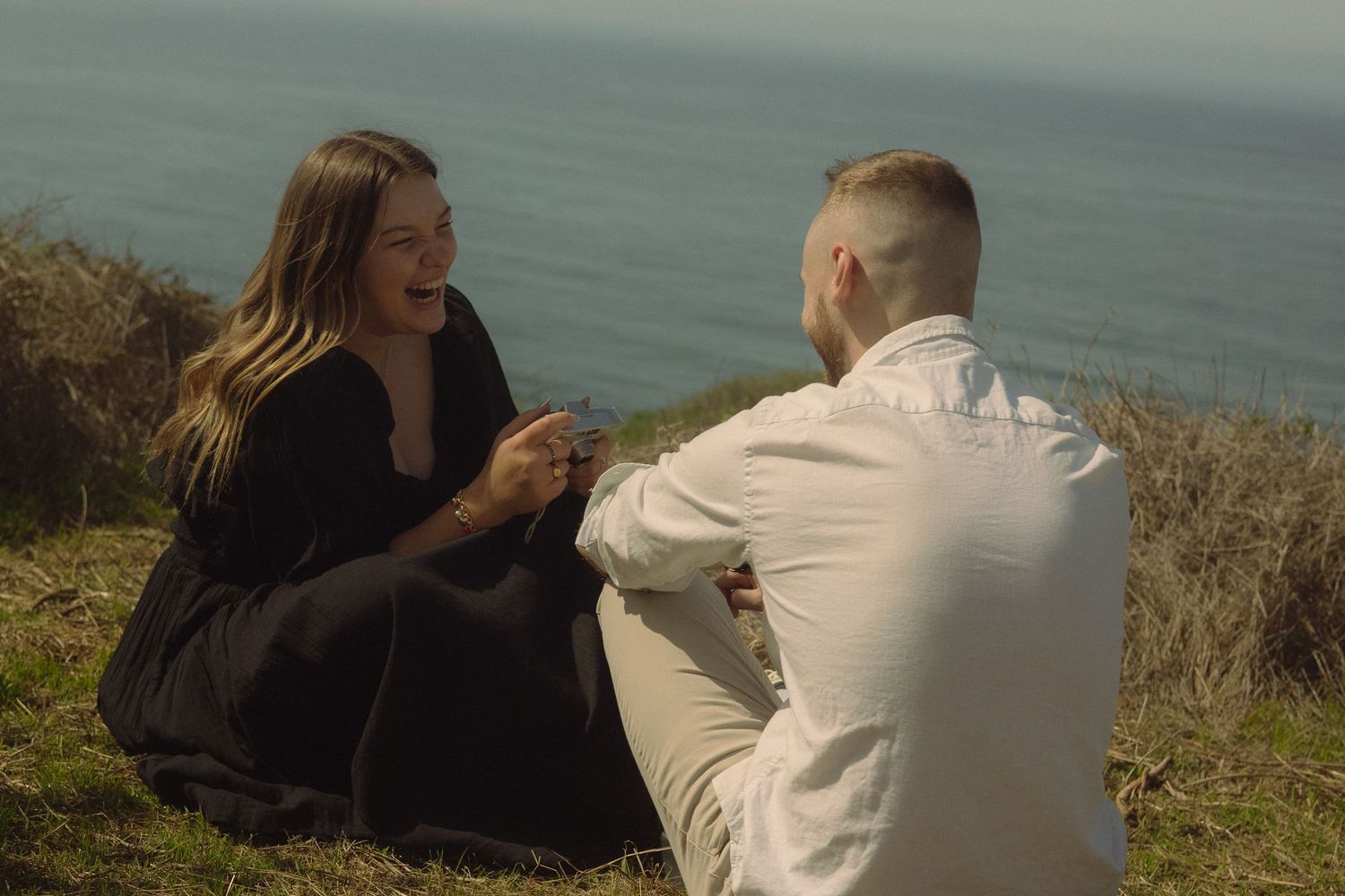 Couple sitting together on grassy oceanside cliff during daytime.