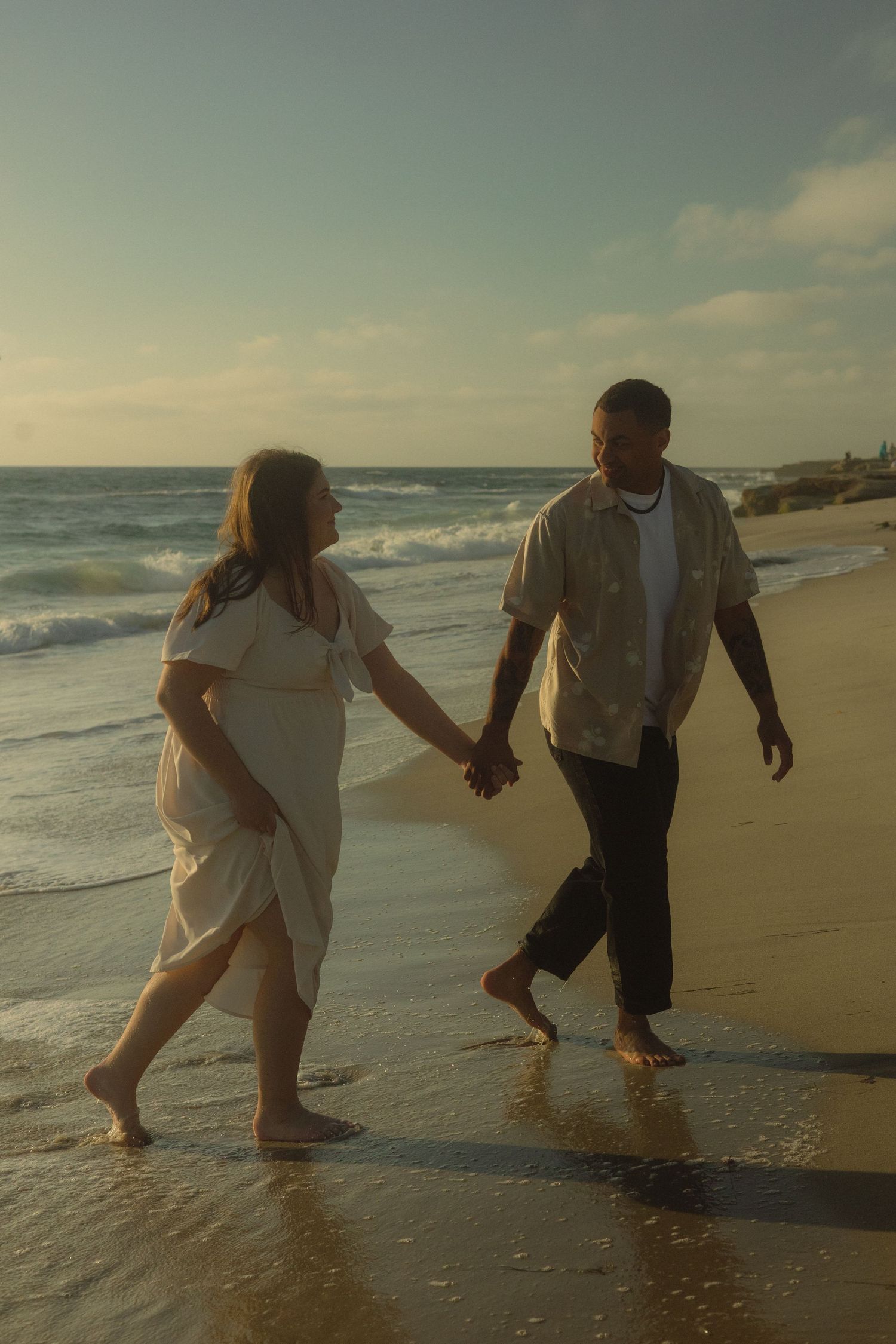 A couple walks hand in hand along the shoreline during golden hour at the beach.