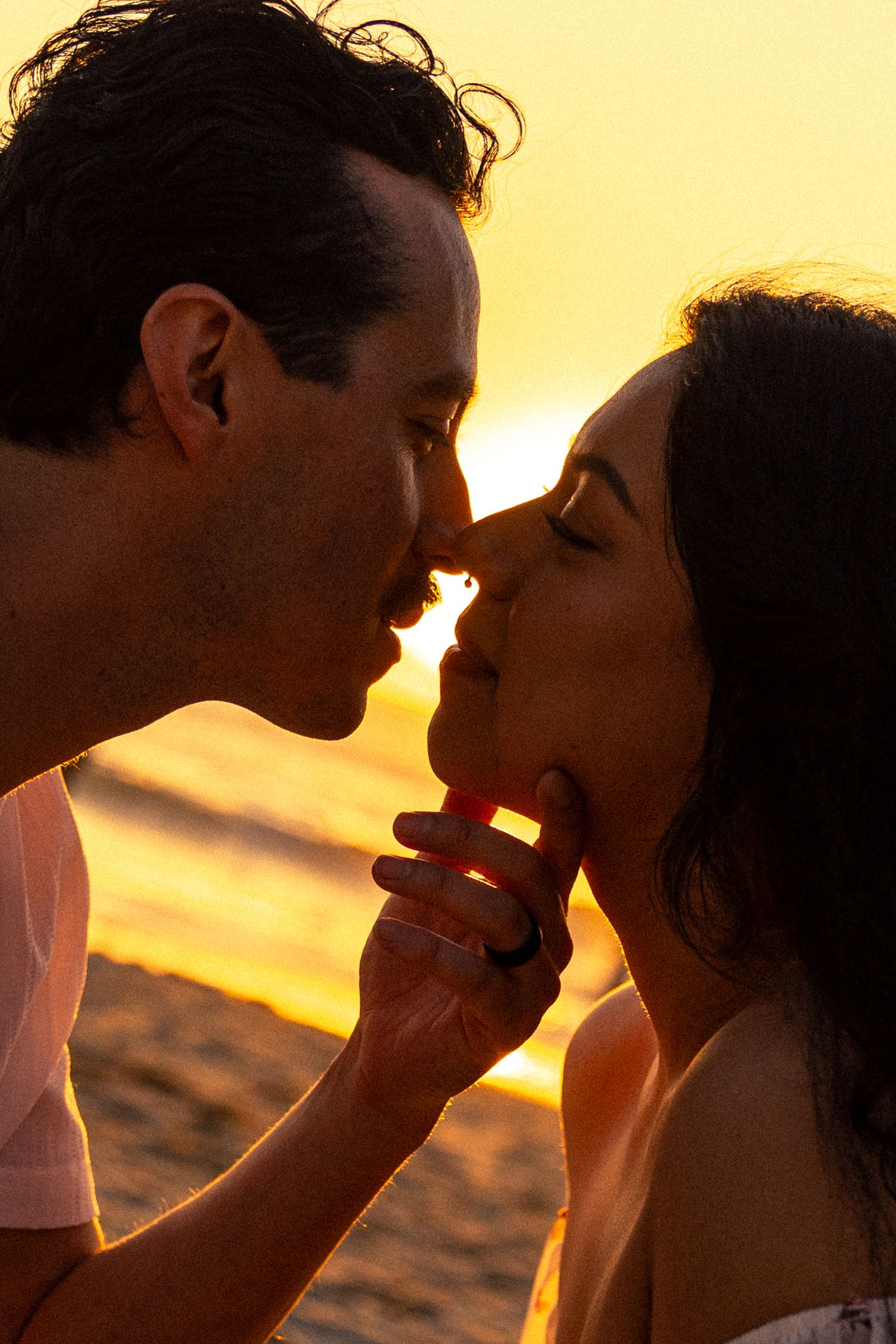 Romantic couple sharing a tender kiss at golden sunset on the beach, silhouetted in warm orange light.