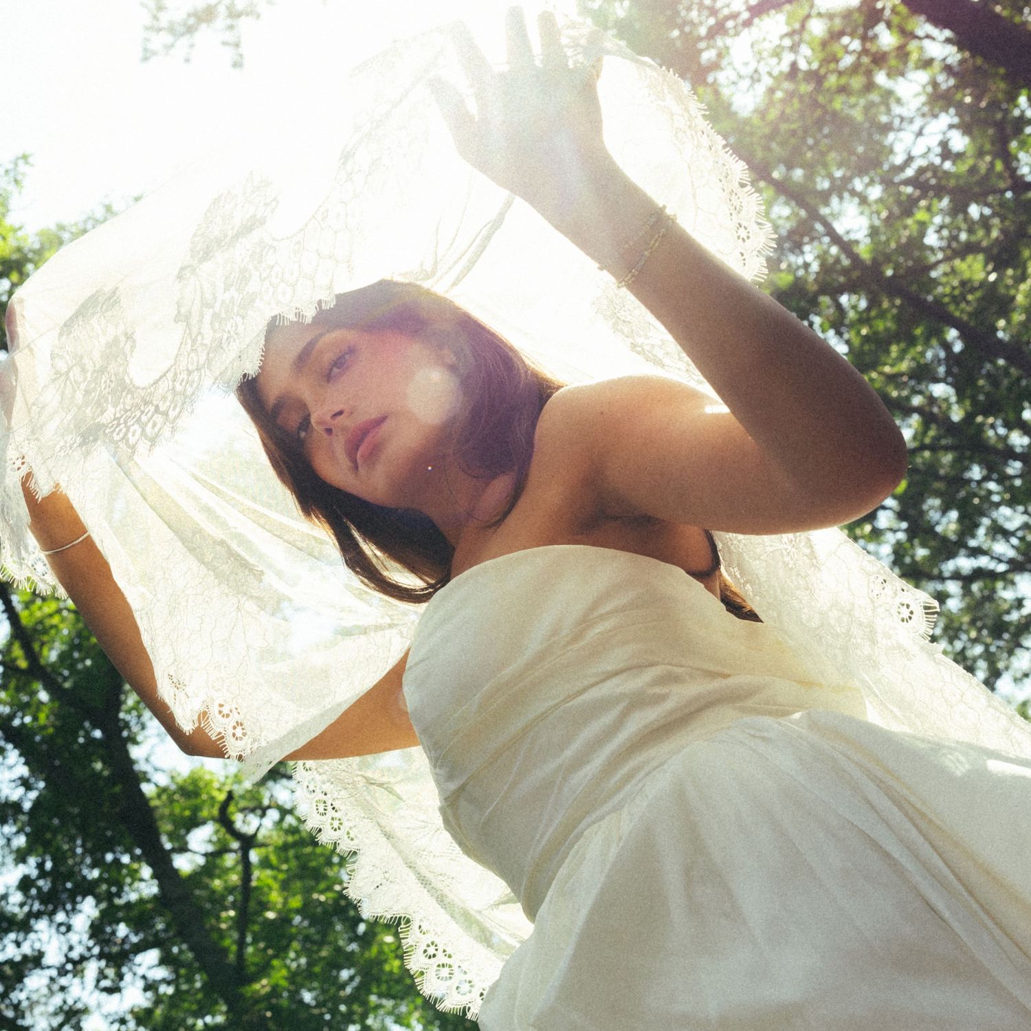 Bride in white lace dress holds veil overhead outdoors, shot from below against sunlit trees.