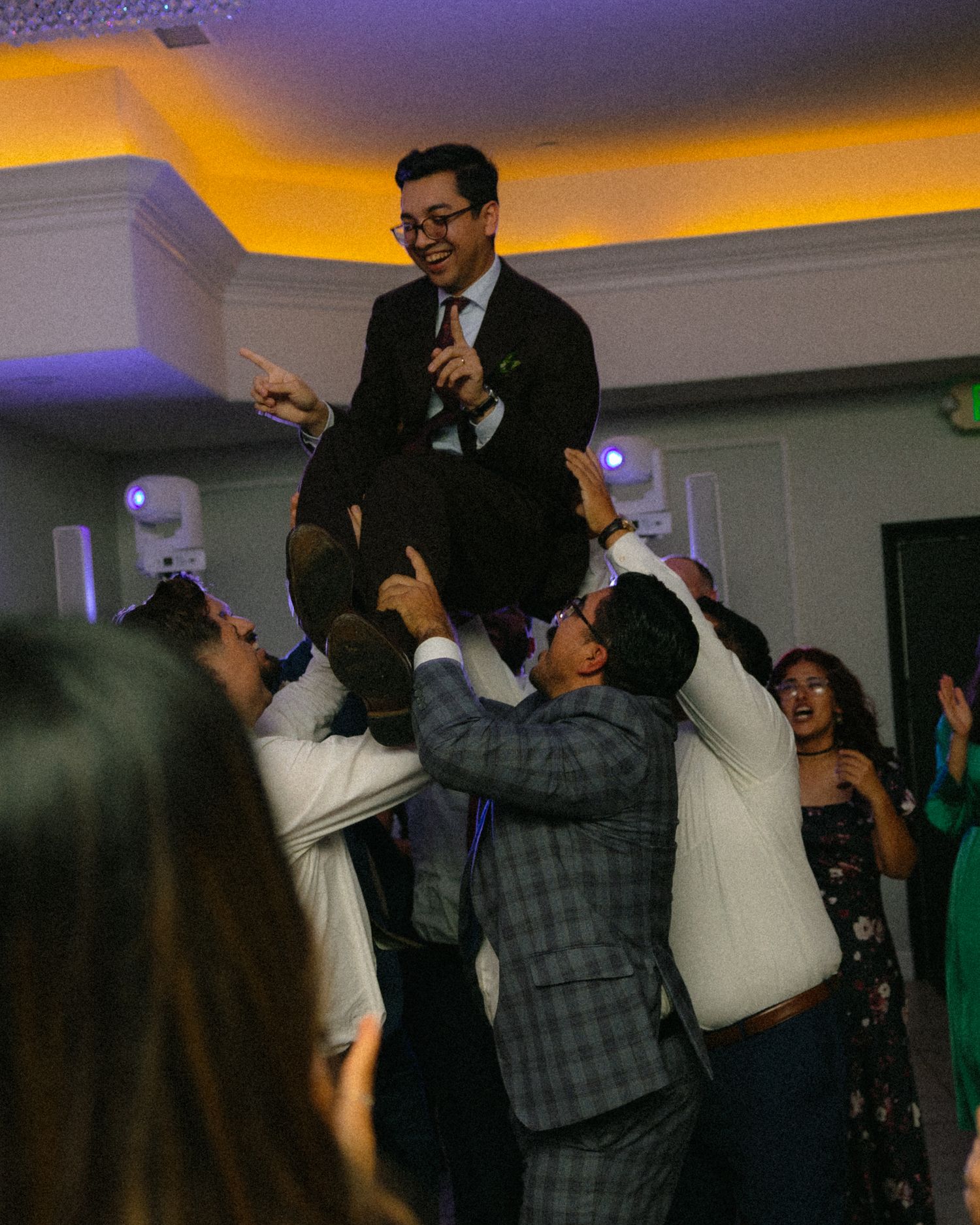People dancing and celebrating at a dimly lit indoor event with yellow and purple ambient lighting.