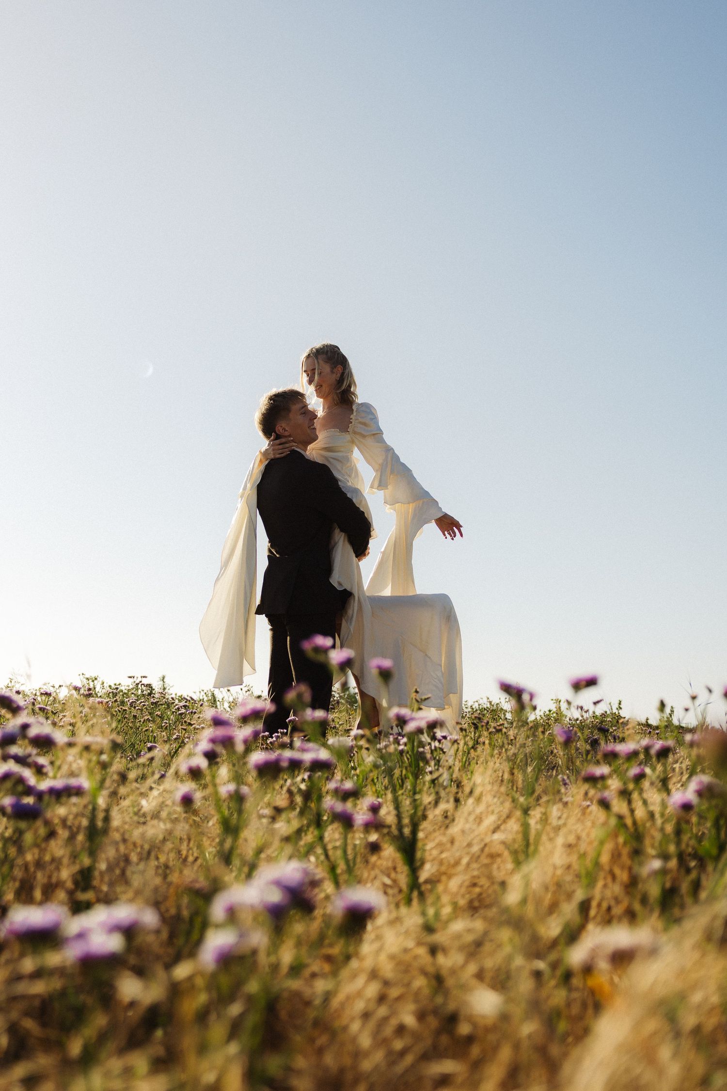 A couple embraces in a field of wildflowers against a blue sky, captured in a series of romantic wedding photos.