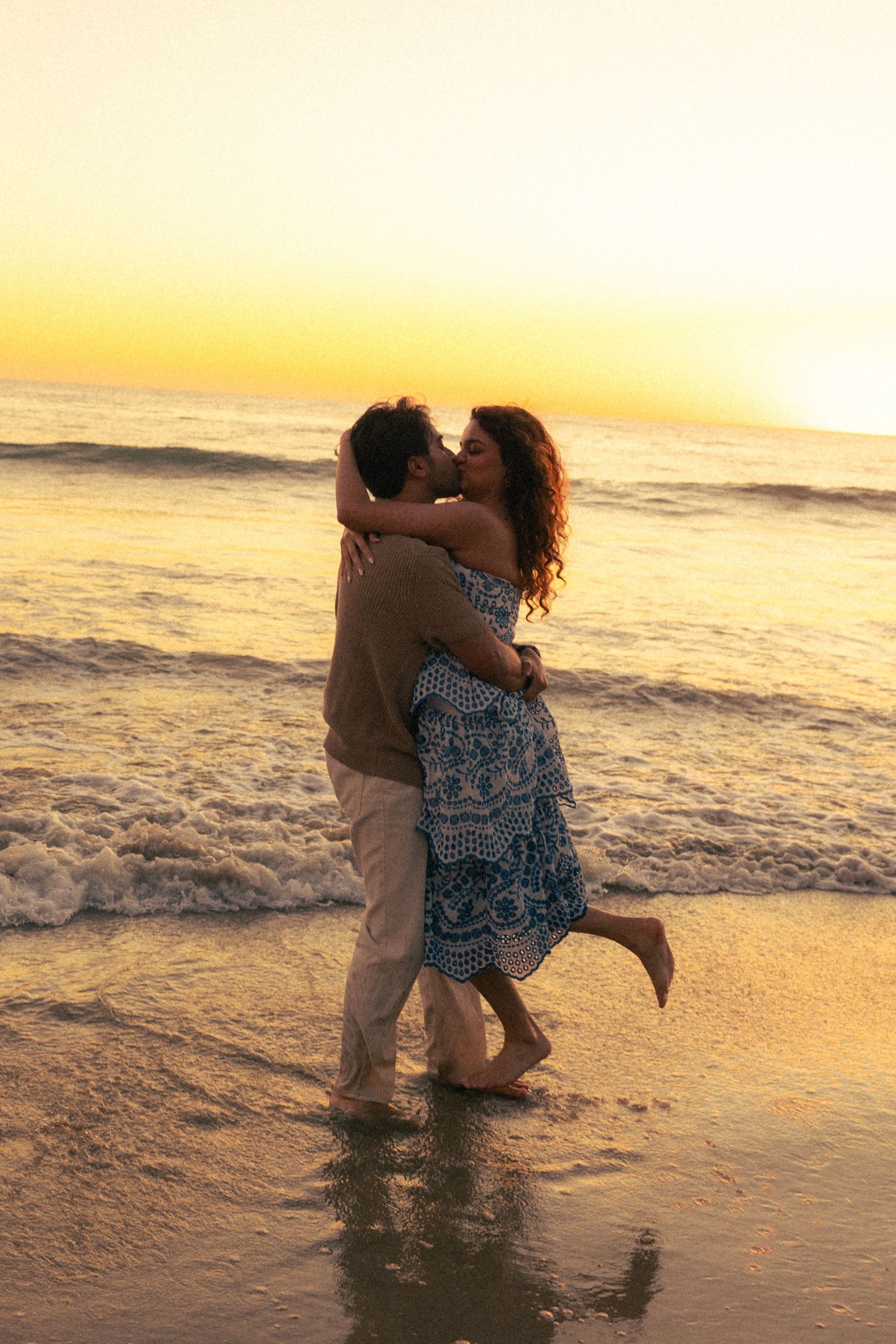 Romantic couple kissing and embracing on a beach at golden sunset, waves washing over their feet.