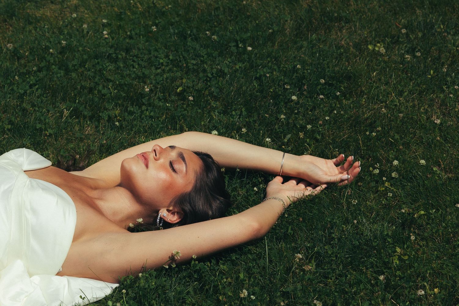Bride in white dress lying on green grass with eyes closed, arms stretched overhead in serene outdoor setting.