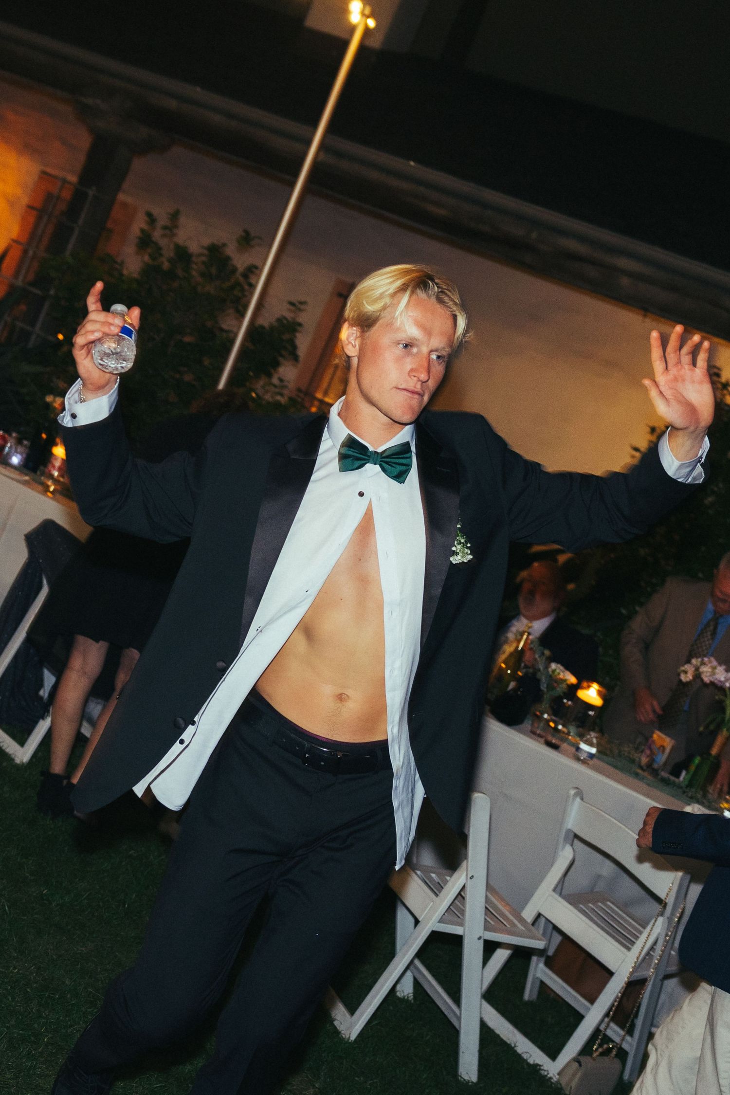 Young blond man in tuxedo with bow tie dancing at formal outdoor evening event holding drink.