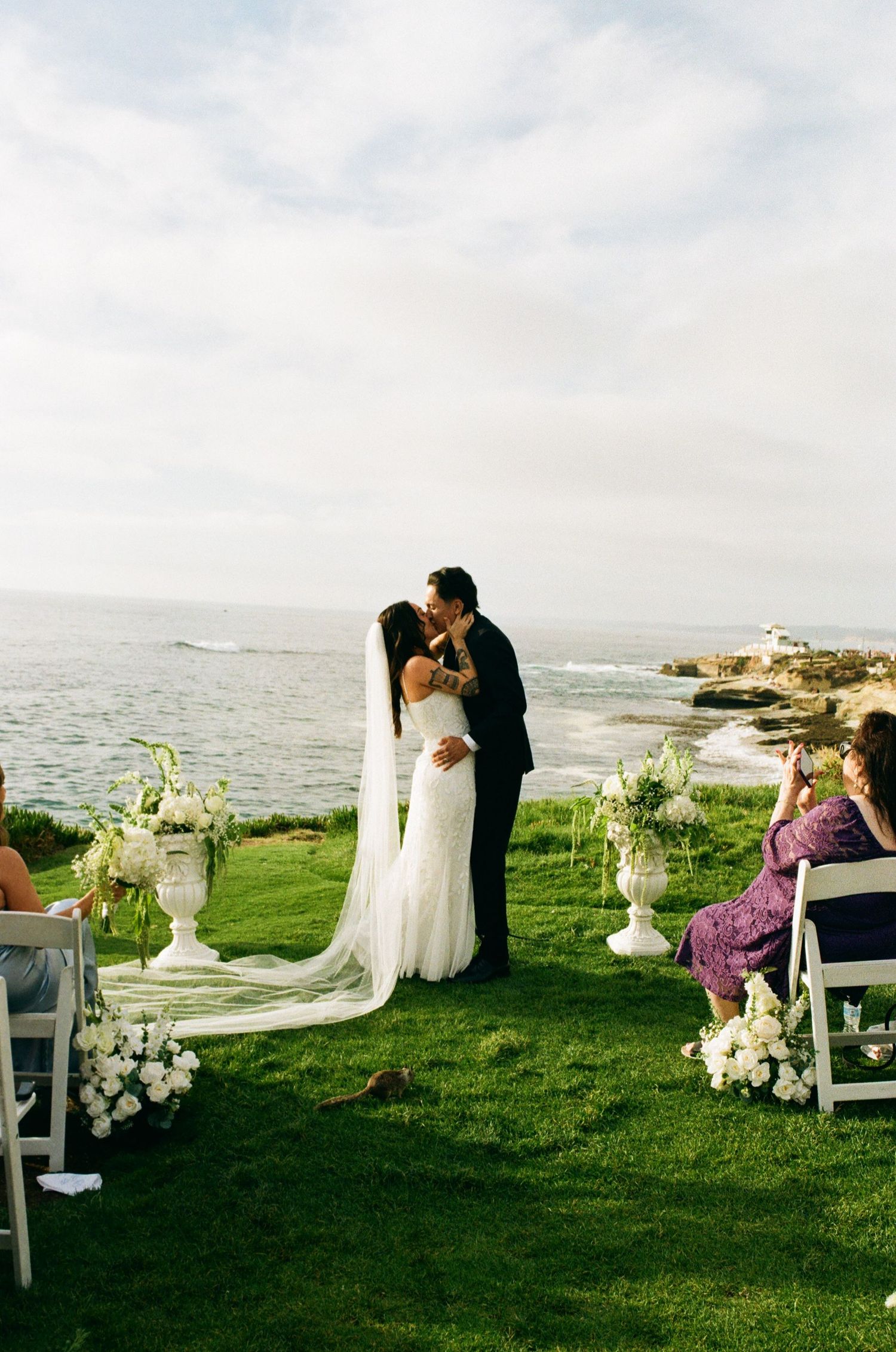 Bride and groom share first kiss at outdoor oceanside wedding ceremony on green lawn with white floral arrangements.