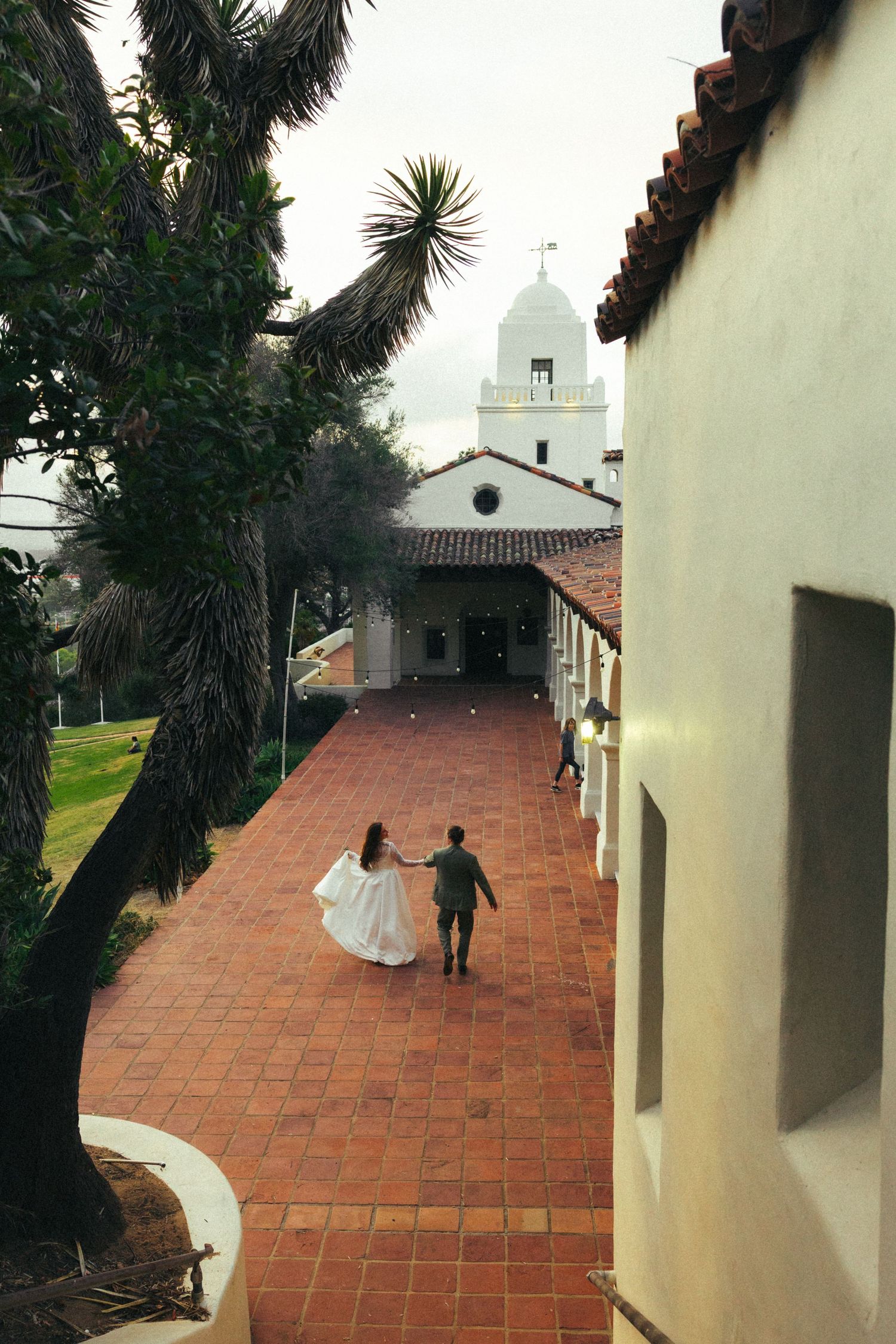 Bride and groom walk hand-in-hand along a terracotta tile courtyard toward a white Spanish colonial chapel.