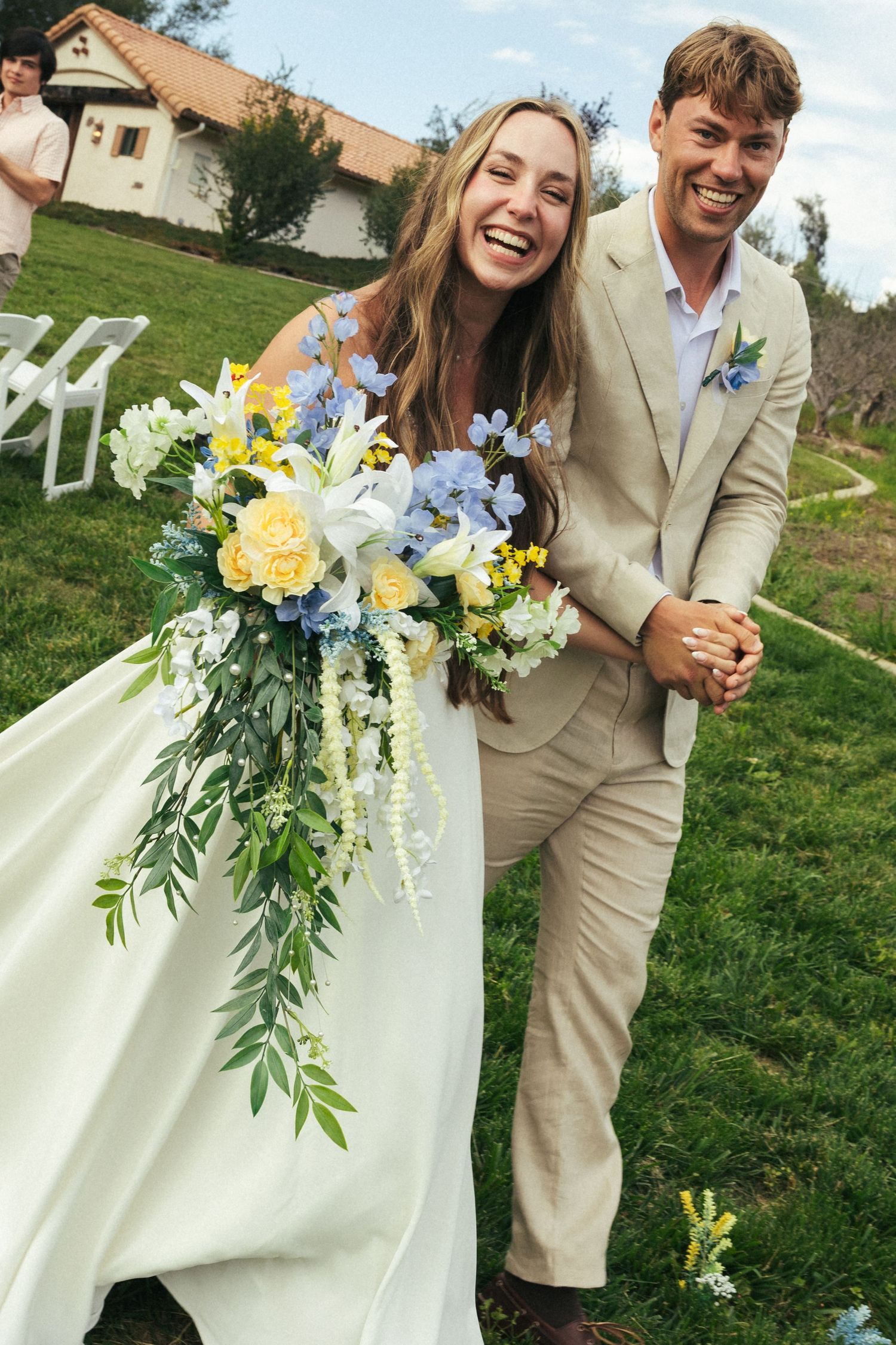 Joyful bride in white gown holding cascading yellow and blue floral bouquet walks with smiling groom in beige suit outdoors.