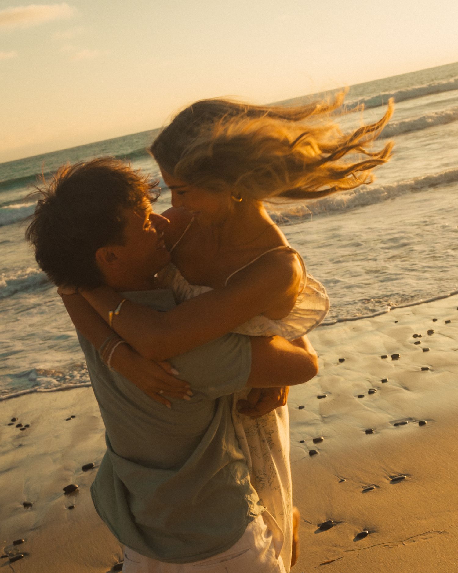 A couple embraces on a beach at sunset with seagulls walking along the shoreline in the golden light.