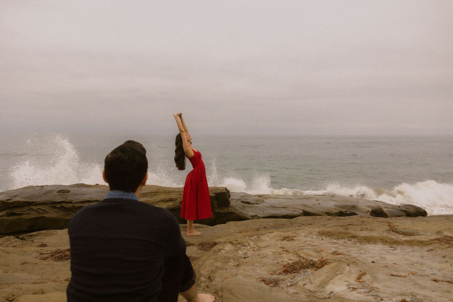 People stand on rocky coast watching large waves crash against shore during stormy weather at the ocean.