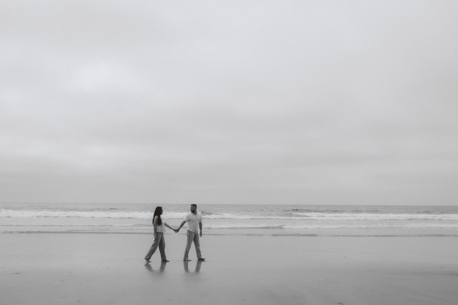 Two silhouettes walk hand in hand on a misty beach shore with gentle waves rolling in the background.