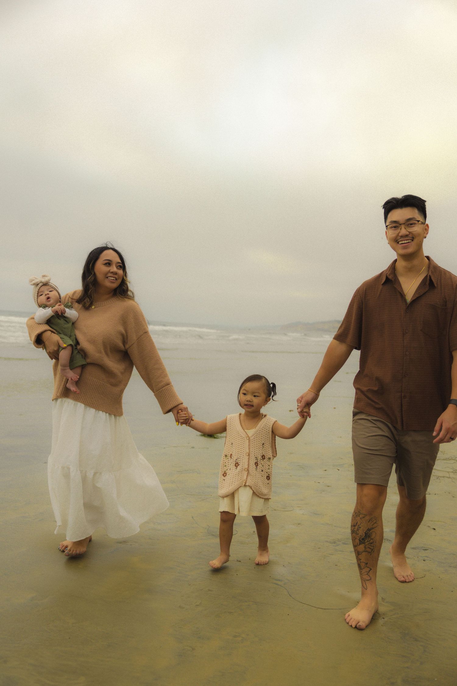 Family walks together barefoot on a foggy beach during golden hour, wearing casual outfits in earth tones.