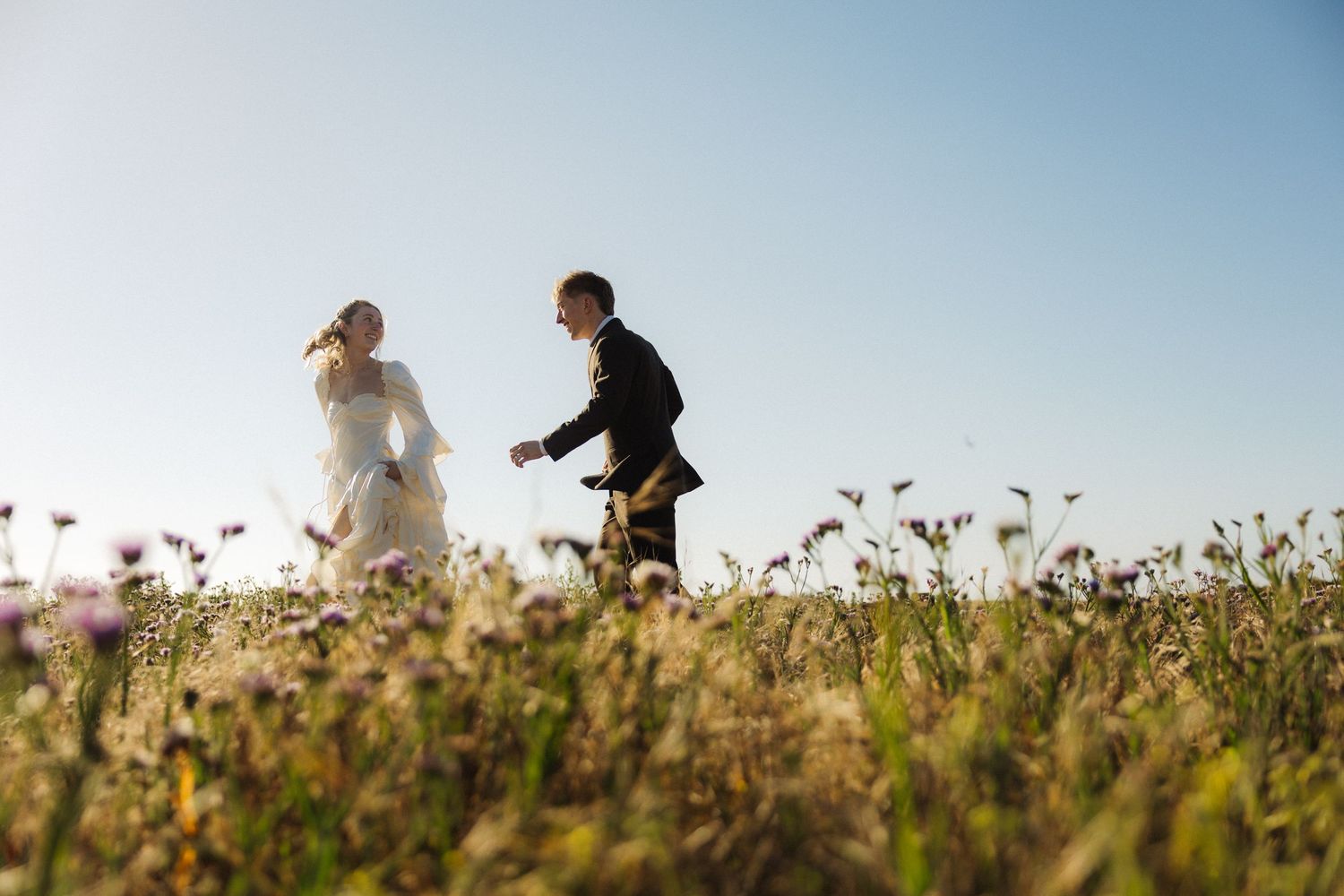 A couple walks through a blooming wildflower field under bright blue skies.
