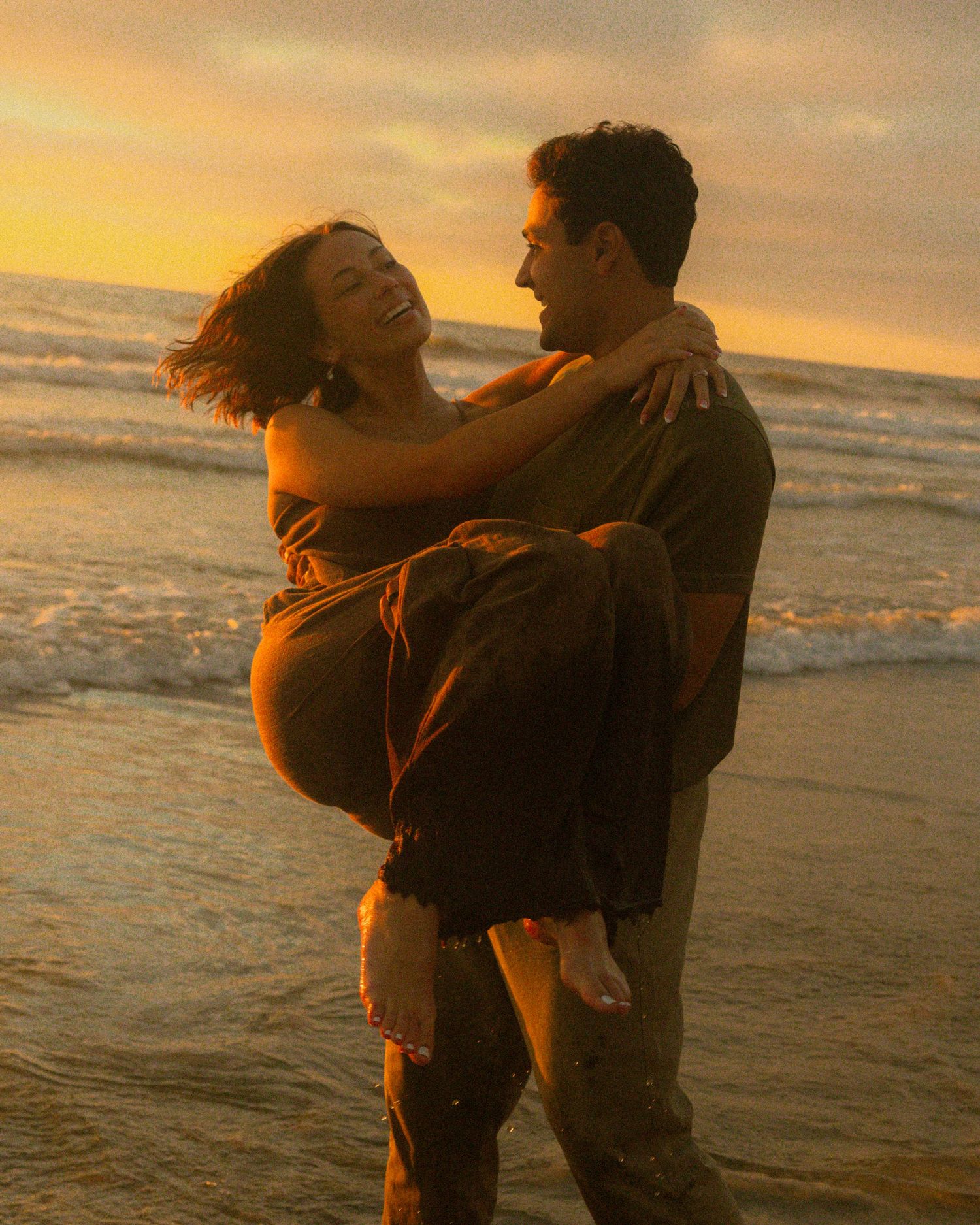 Couple embracing on a beach during golden hour with waves and warm sunlight creating a romantic atmosphere.