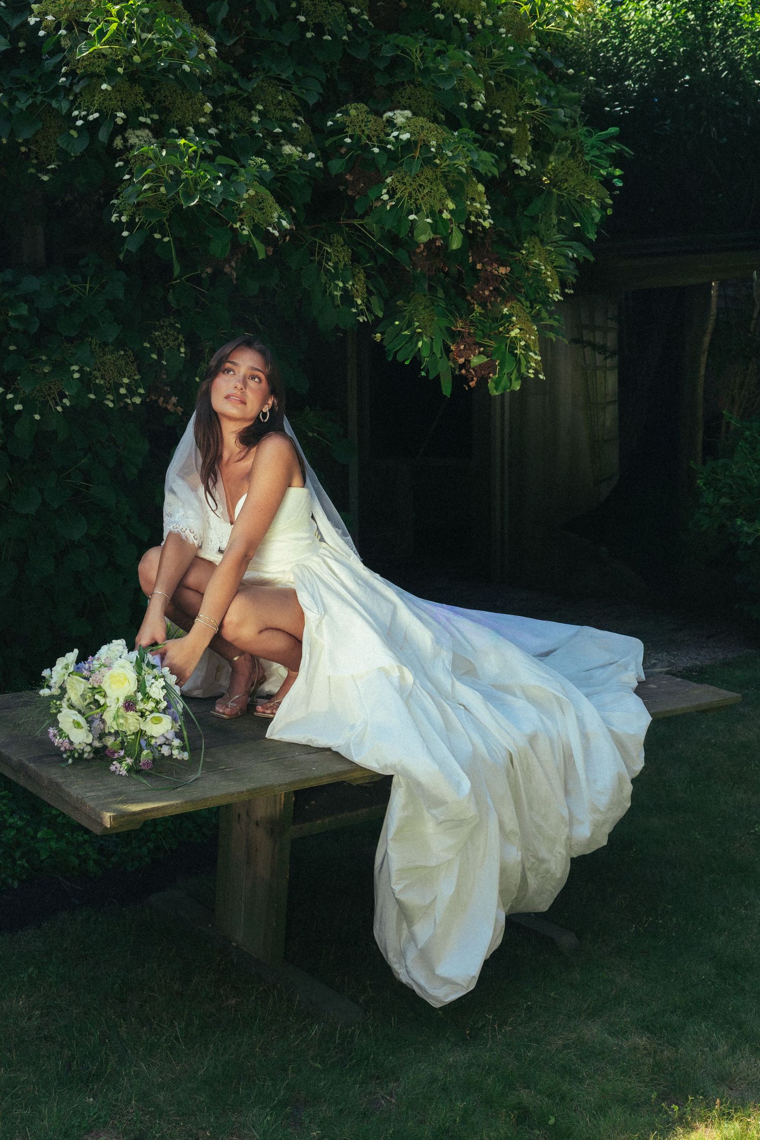 Bride in white gown sitting on wooden bench with bouquet in lush garden.