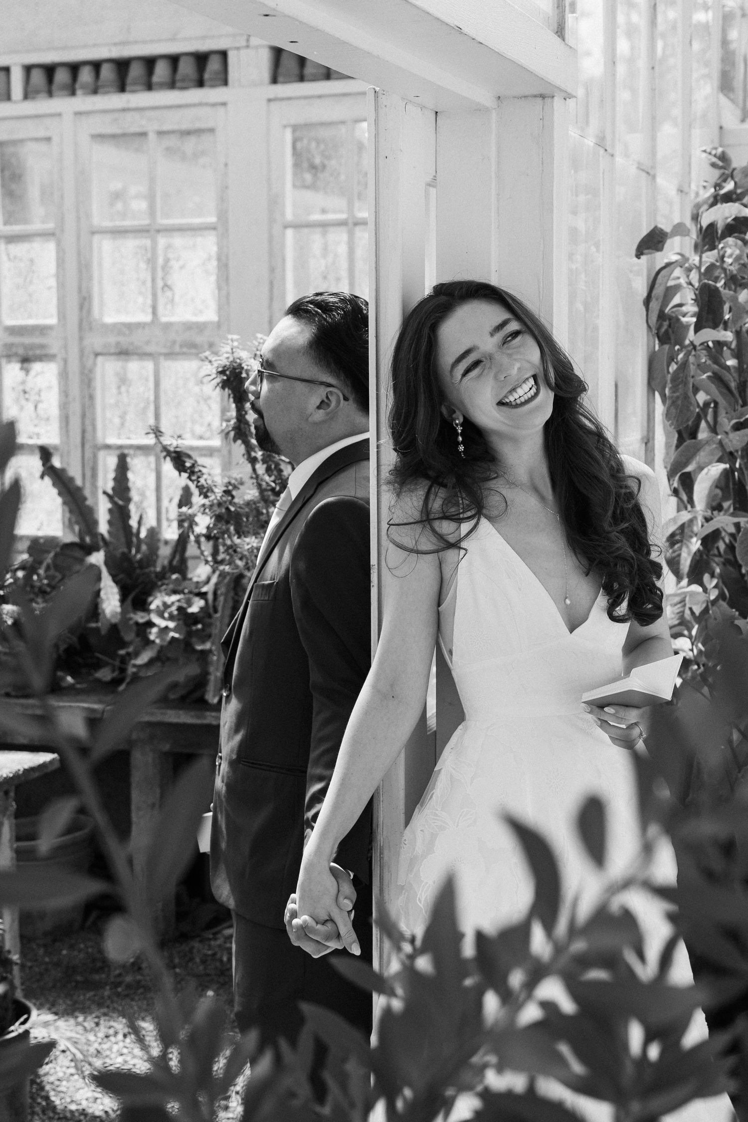 Black and white photo of a laughing bride in a white gown holding hands with a suited groom near lush greenery.