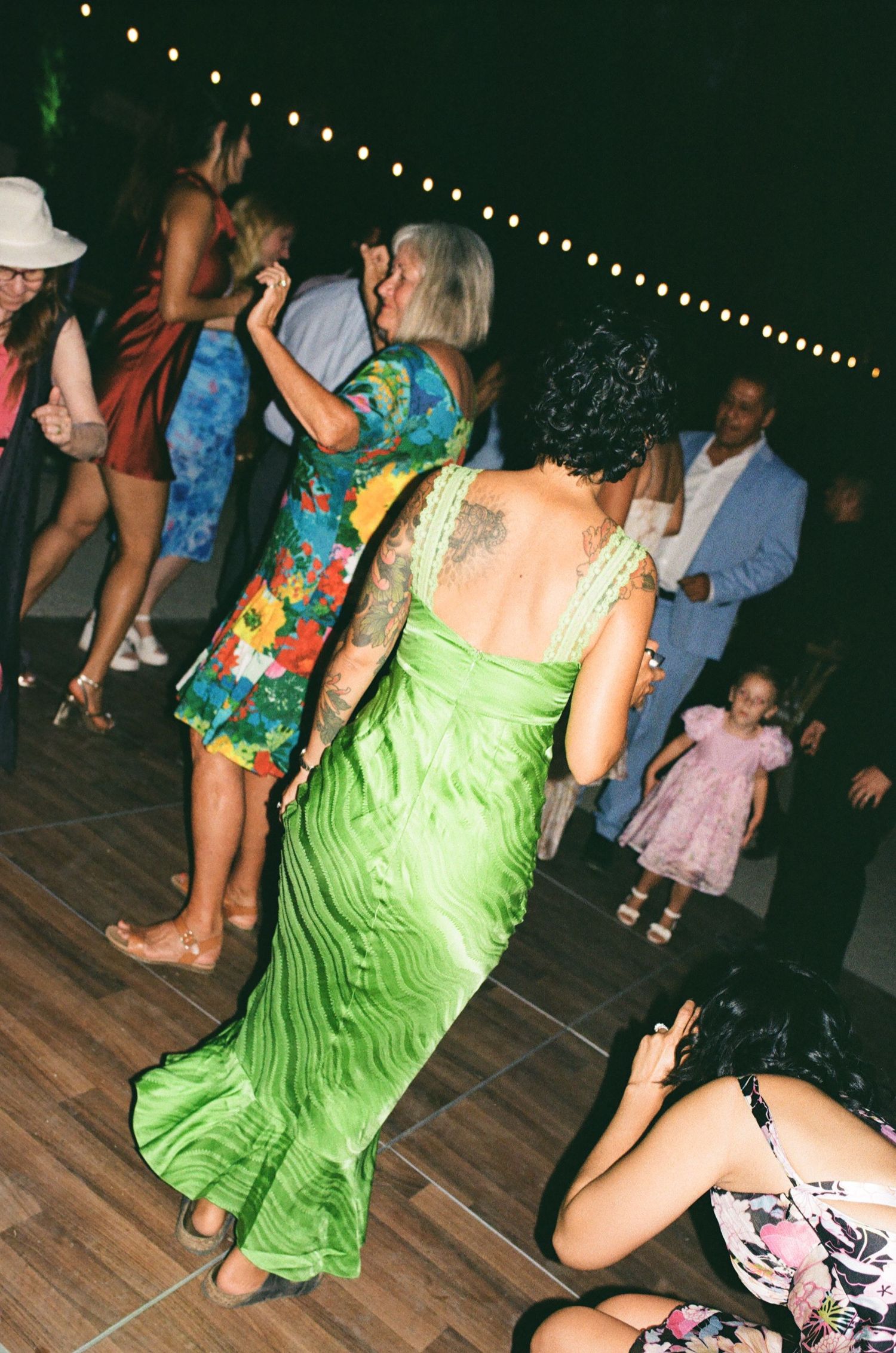 Woman in green maxi dress with back tattoo dancing at an outdoor evening party with string lights.