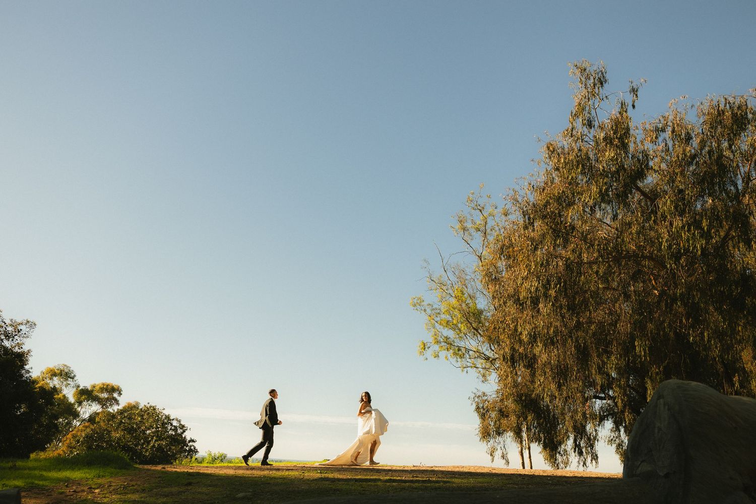 Couple walking together on a hilltop at sunset with golden trees silhouetted against a clear blue sky.