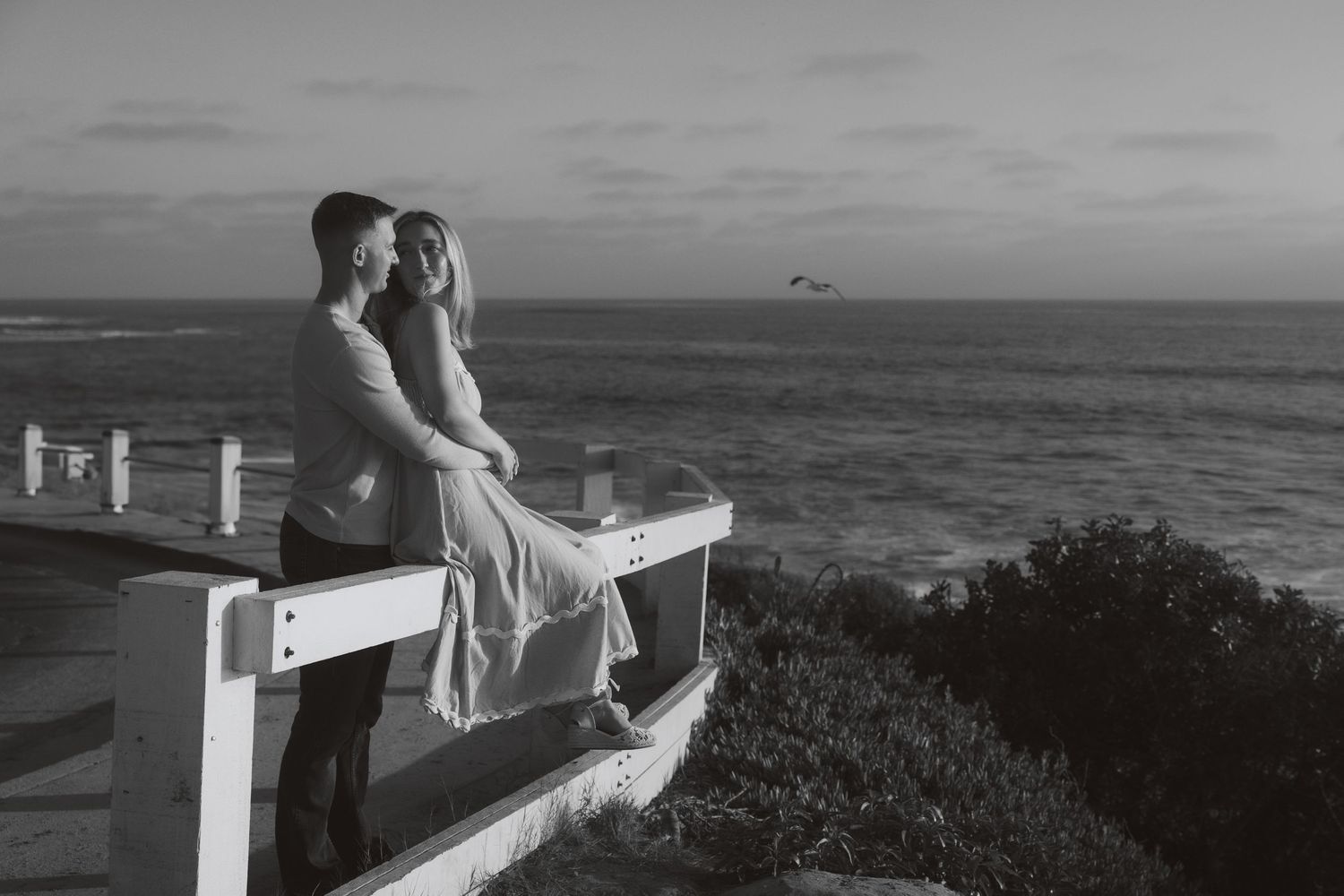 A black and white photo shows people relaxing on a coastal deck overlooking the ocean on a sunny day.