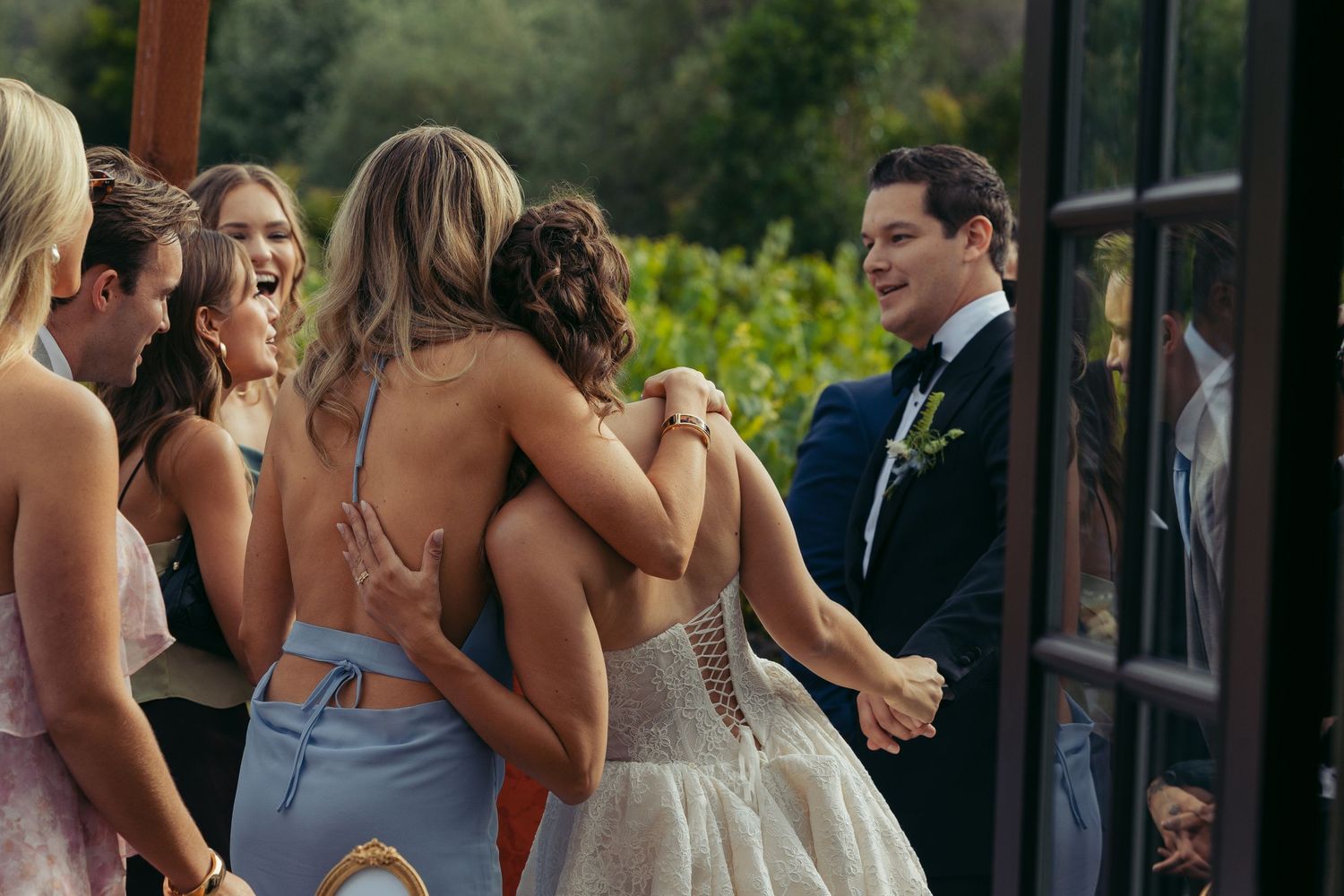 Bride in lace gown embraced by bridesmaids in blue dresses at outdoor vineyard wedding celebration.