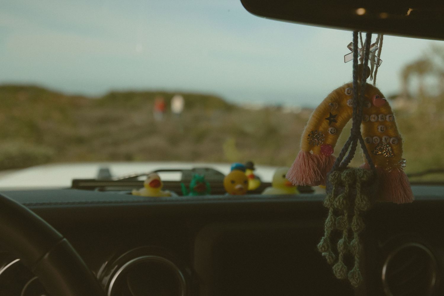 A decorative hanging ornament dangles from a car rearview mirror against a blurred dashboard.