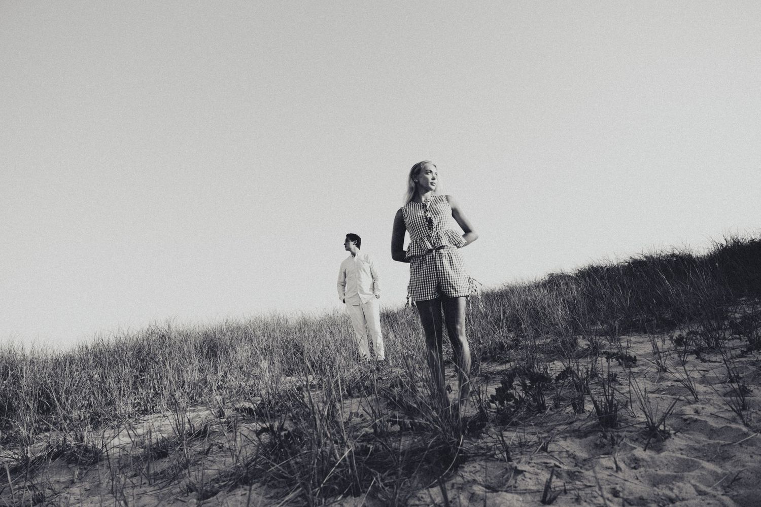 Two people walking on sandy dunes with sparse grass in a black and white coastal landscape photo.