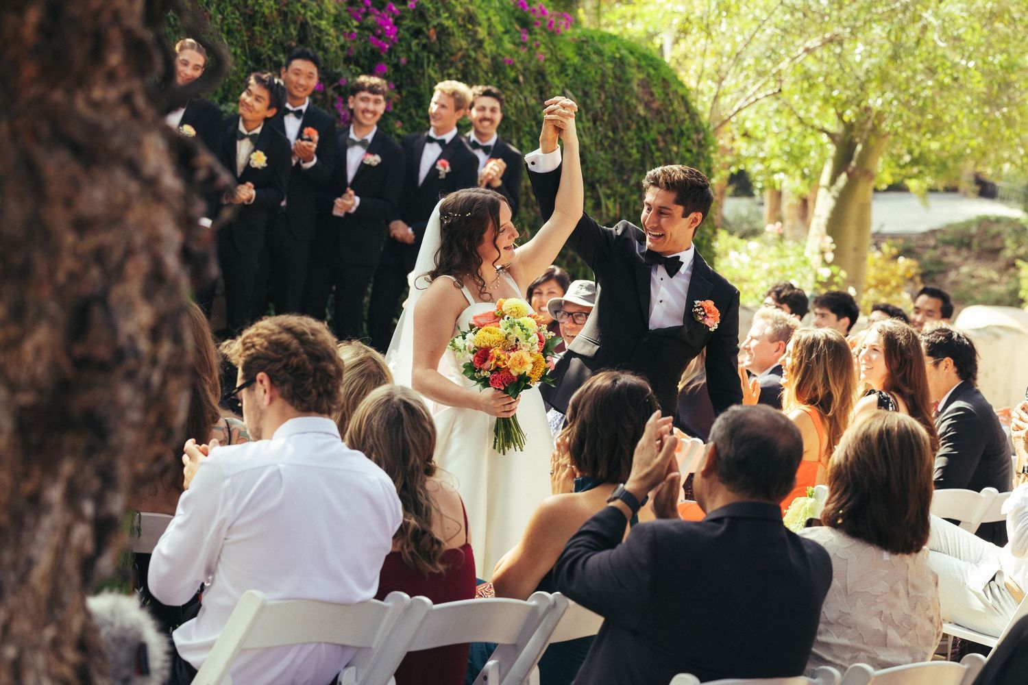 Joyful newlyweds raise hands at outdoor wedding ceremony surrounded by cheering guests and groomsmen in black tuxedos.