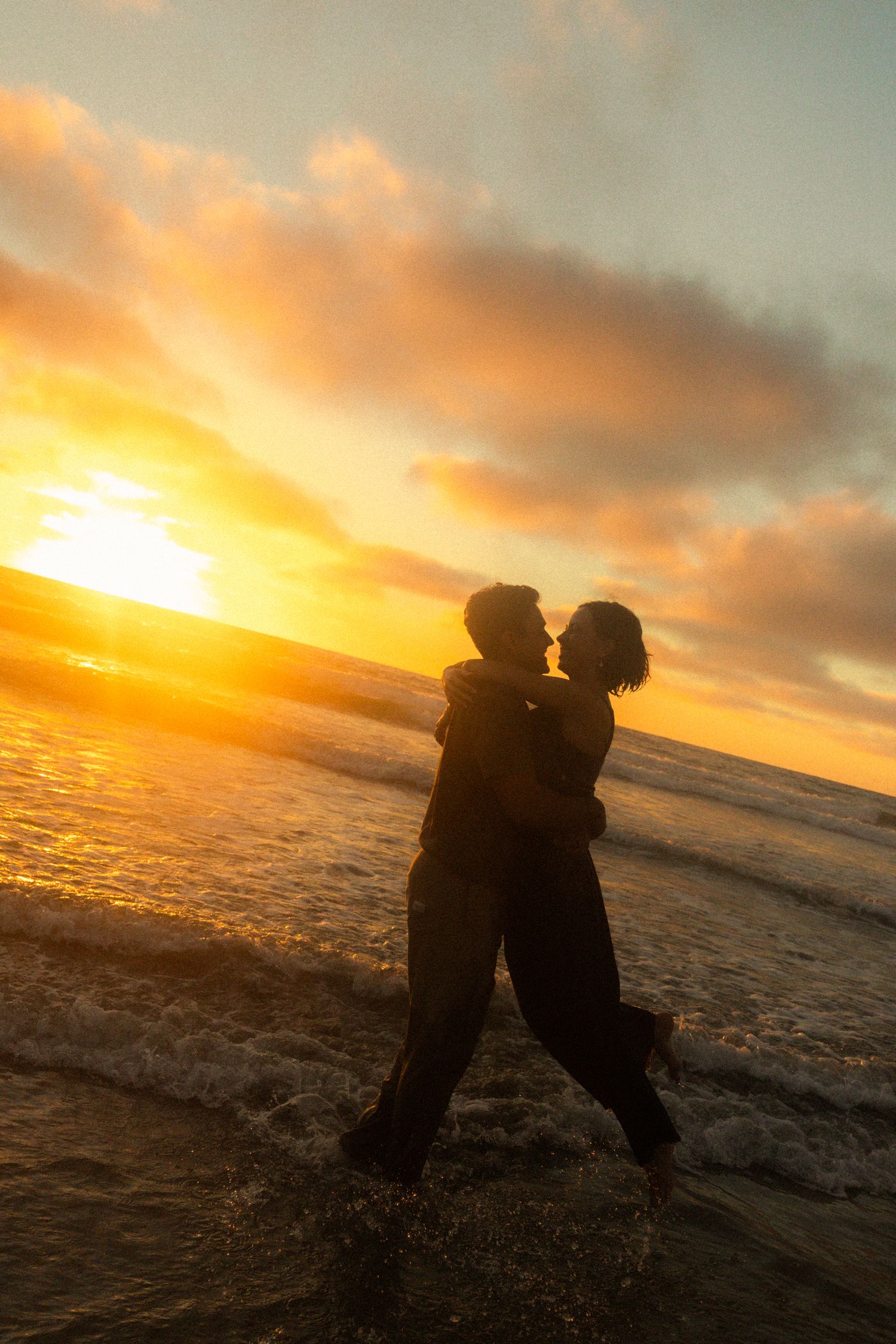Silhouettes of two figures sitting close together on a beach during a dramatic orange sunset.