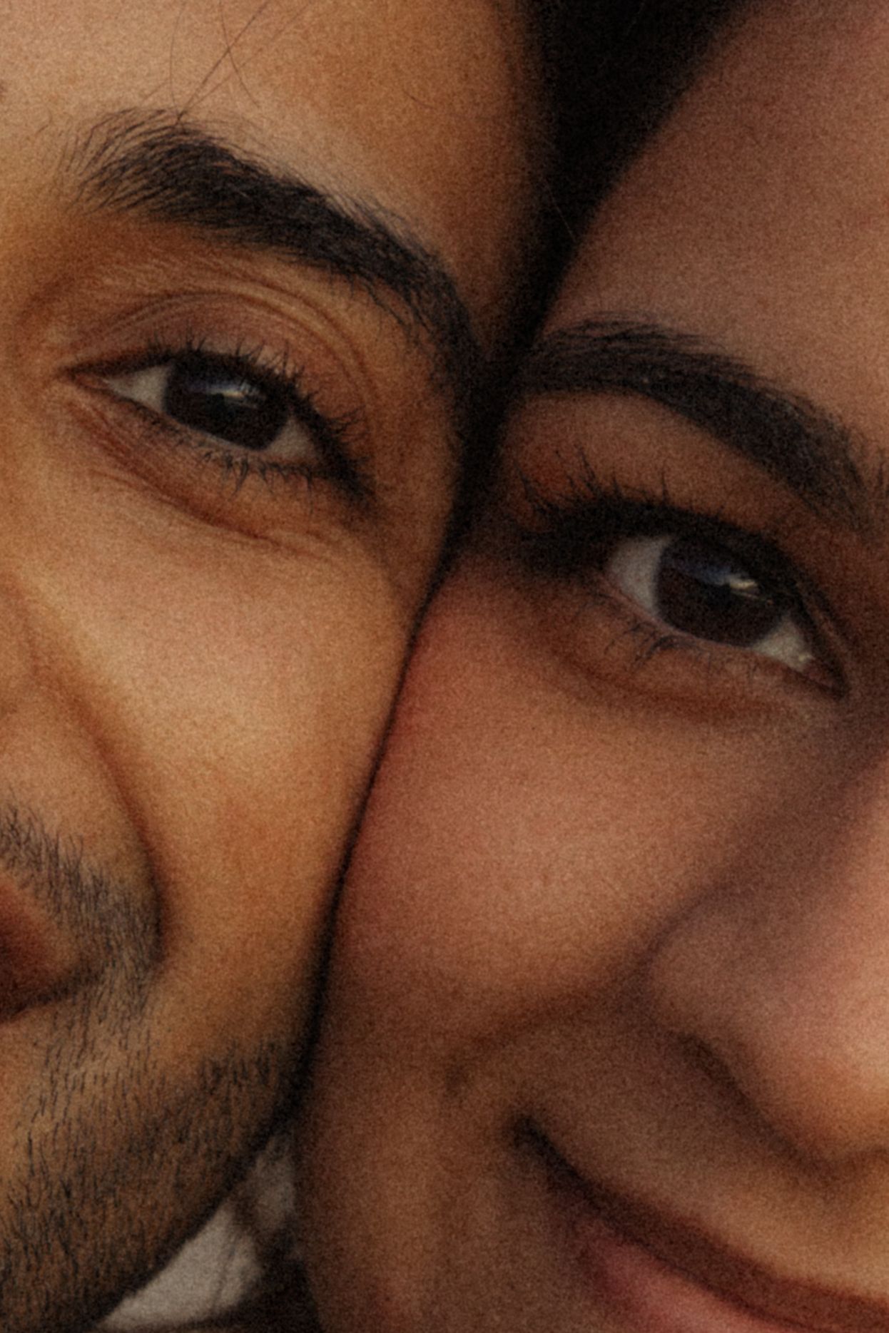 Close-up portrait of two people cheek to cheek, smiling warmly with dark eyes and brows visible.