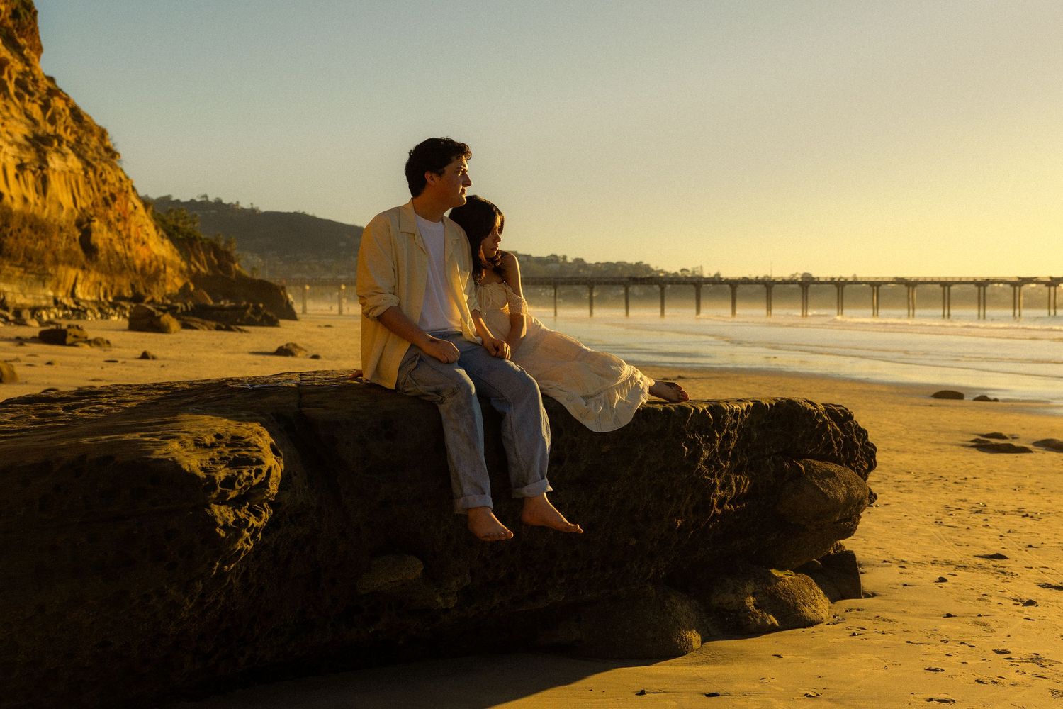 A couple sits together on a large rock at sunset on a beach with a wooden pier in the background.