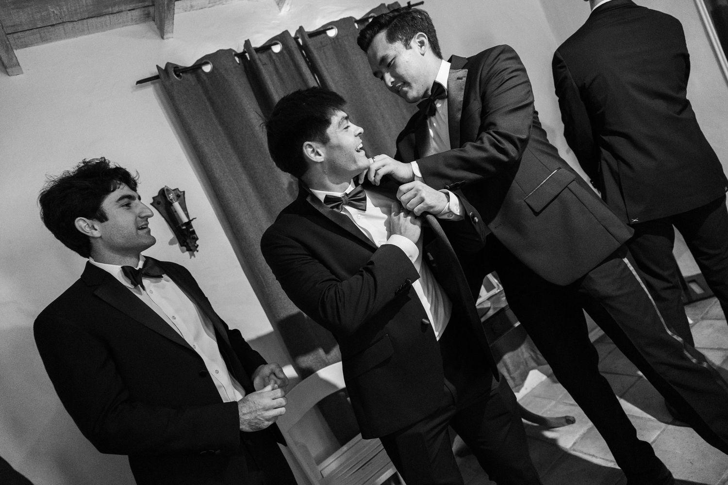 Three men in tuxedos help each other adjust bow ties before a formal event.