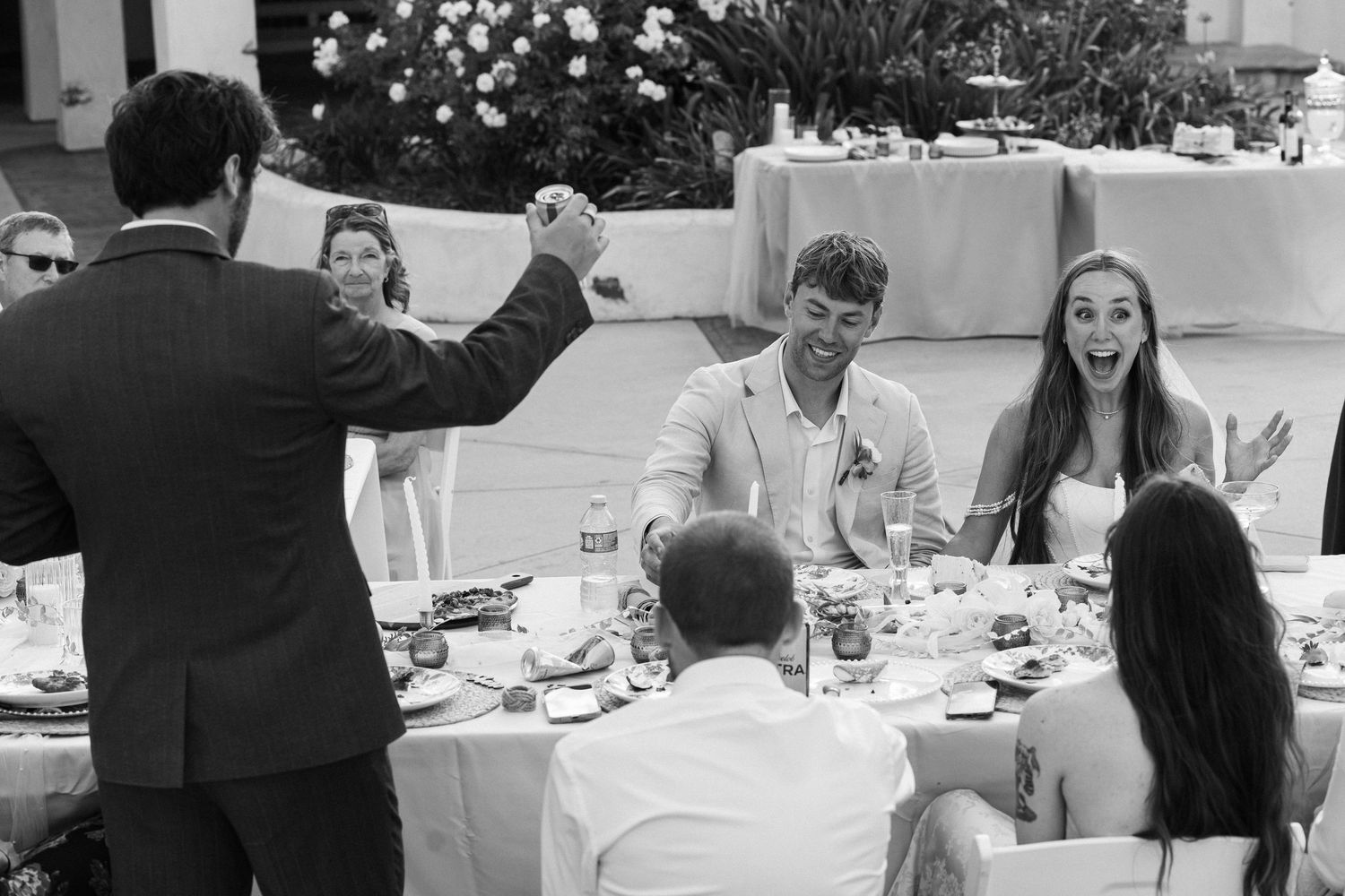 Black and white wedding reception photo of guests laughing and celebrating at an outdoor dinner table.