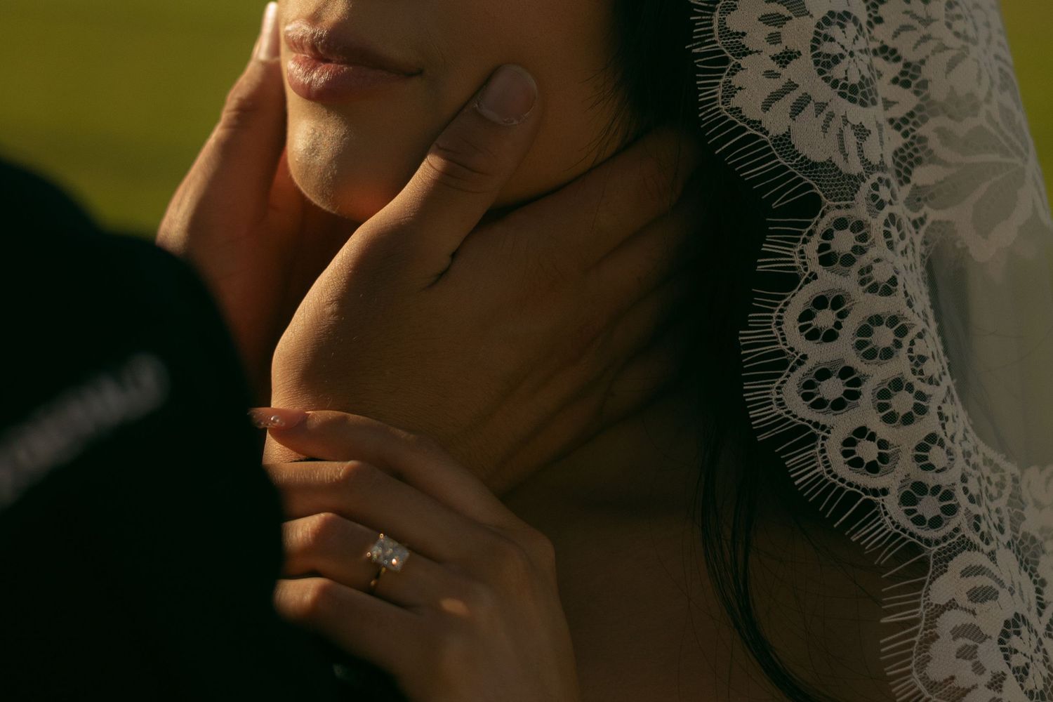 Close-up shot of hands with a delicate engagement ring against white lace in warm golden light.