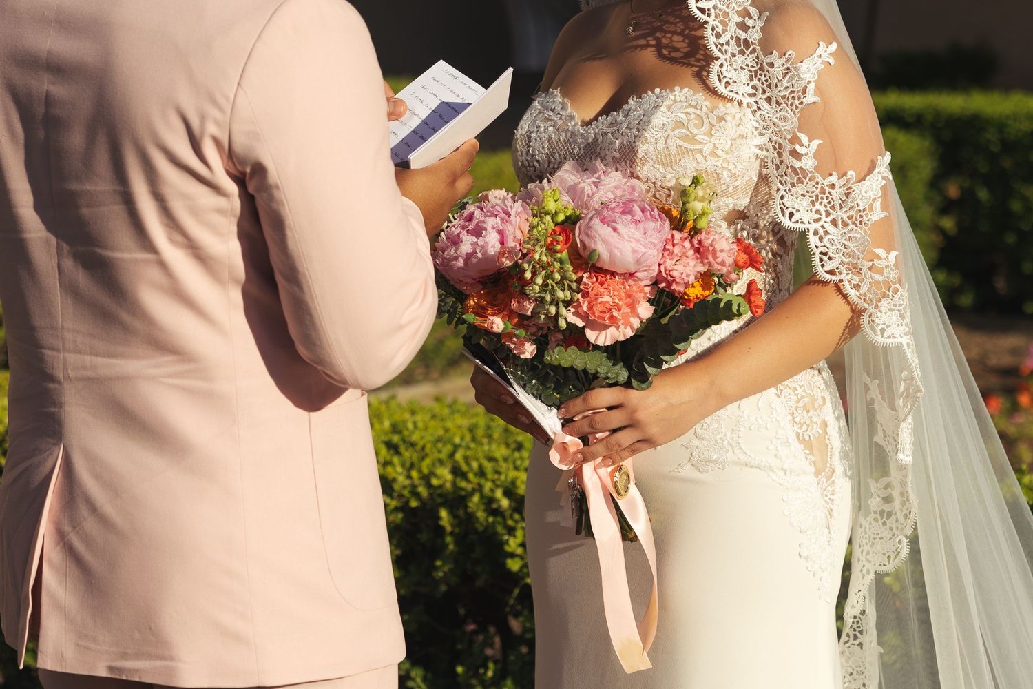 Bride in lace wedding dress holding colorful pink peony bouquet while groom in blush suit reads vows outdoors.