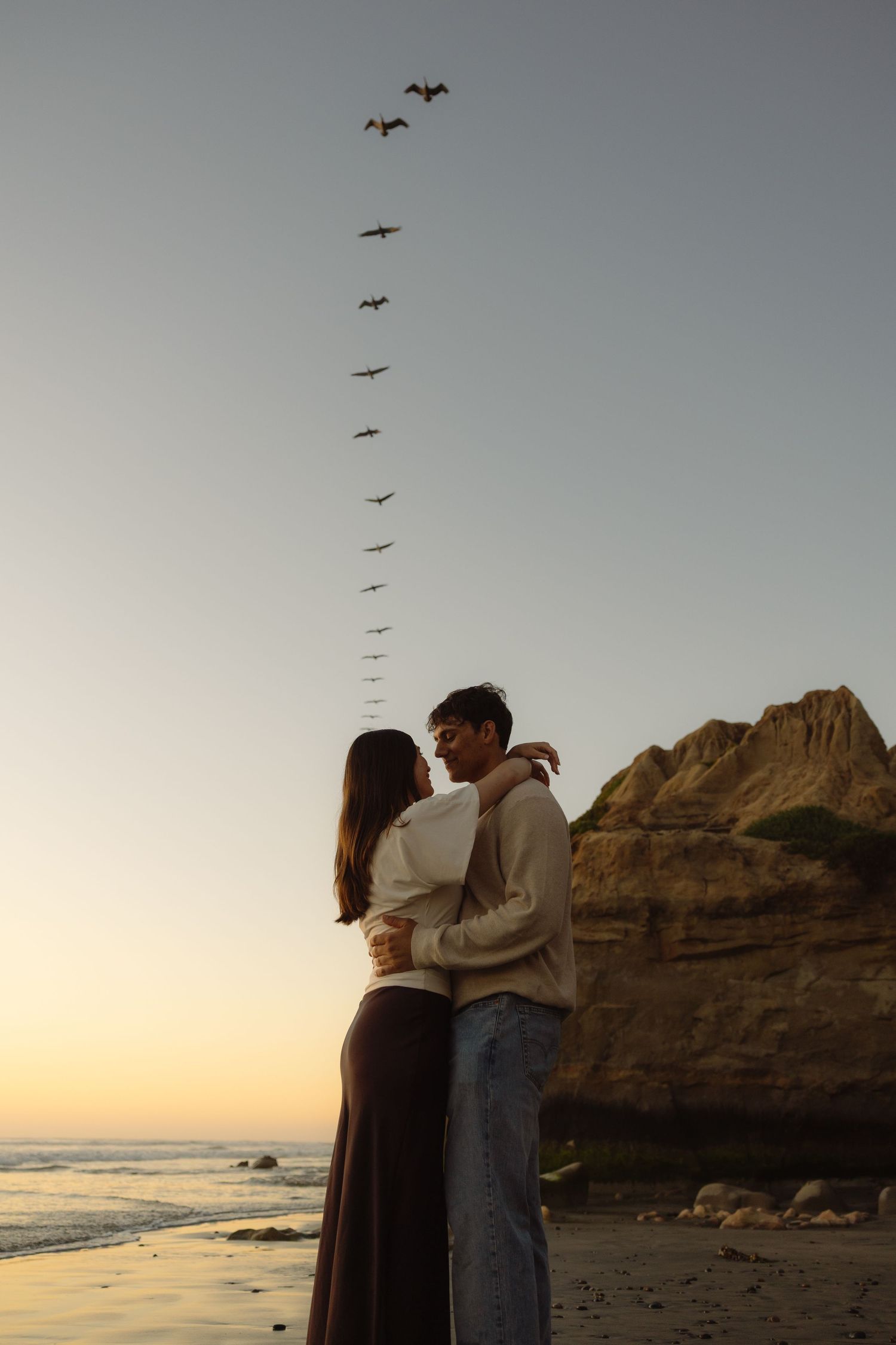 A couple embraces on a beach at sunset with birds flying overhead and coastal cliffs in the background.