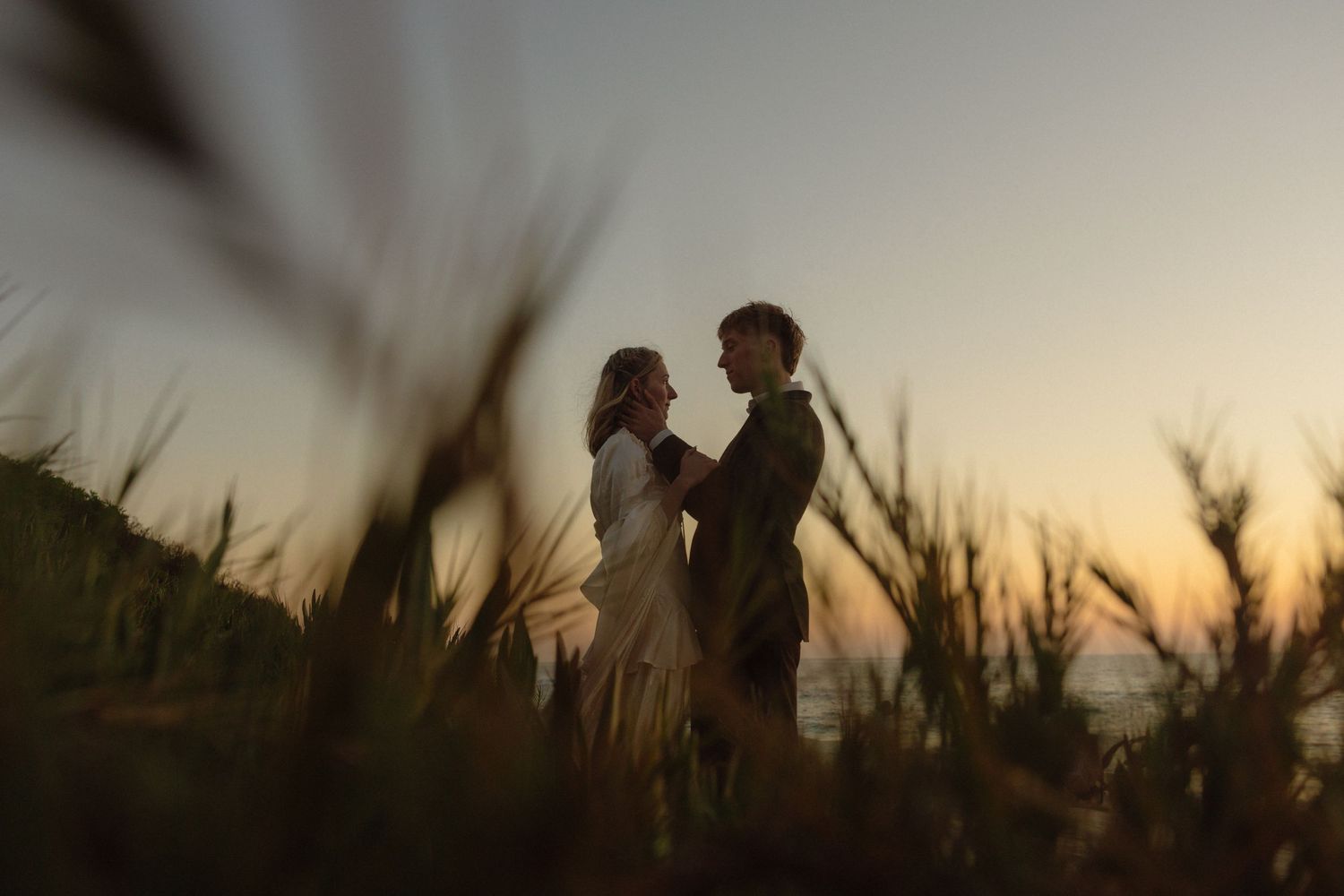 Silhouettes of a couple against a sunset sky with tall grass in the foreground create a romantic scene.