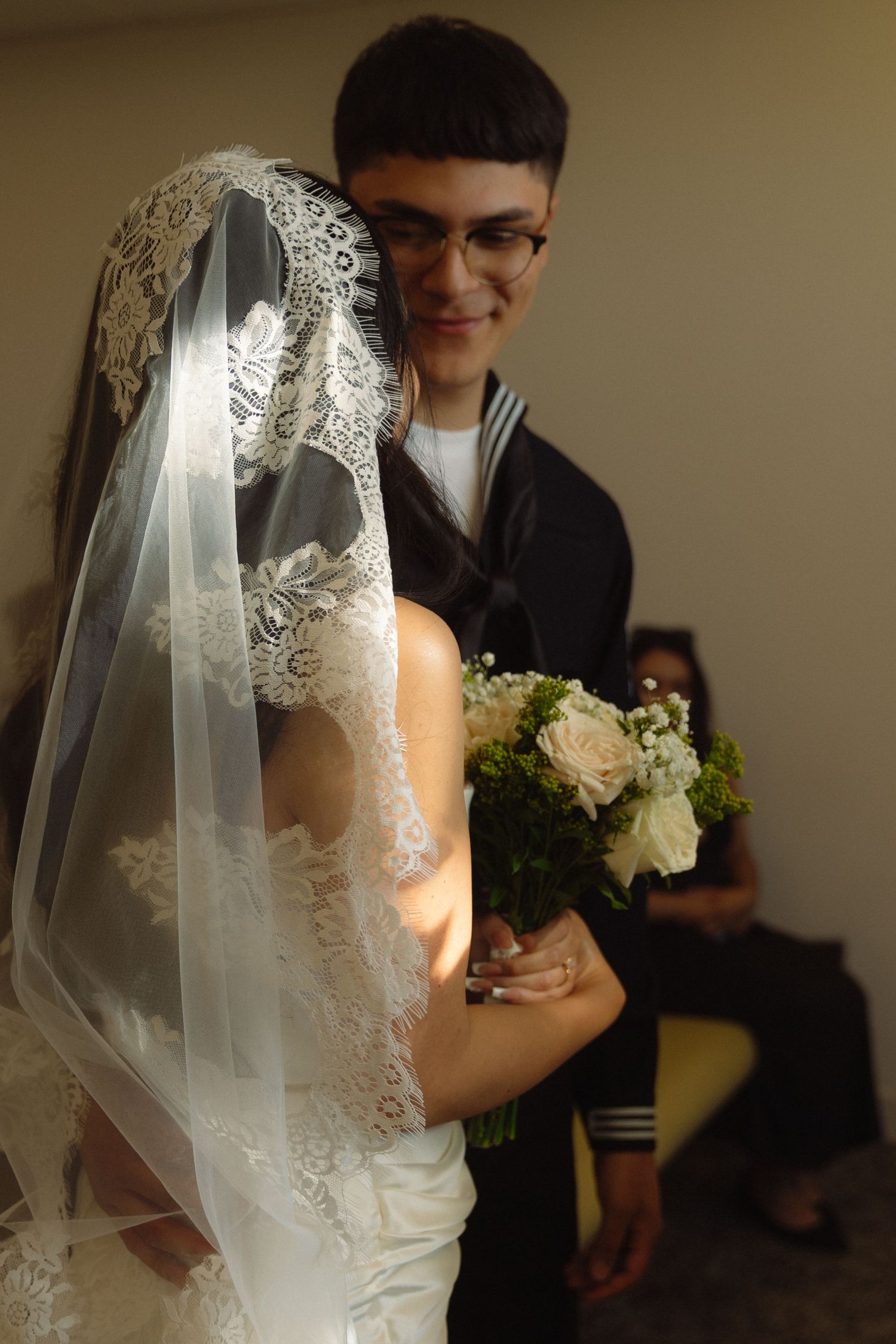 A bride in a delicate lace veil holds a white rose bouquet during an intimate wedding moment.
