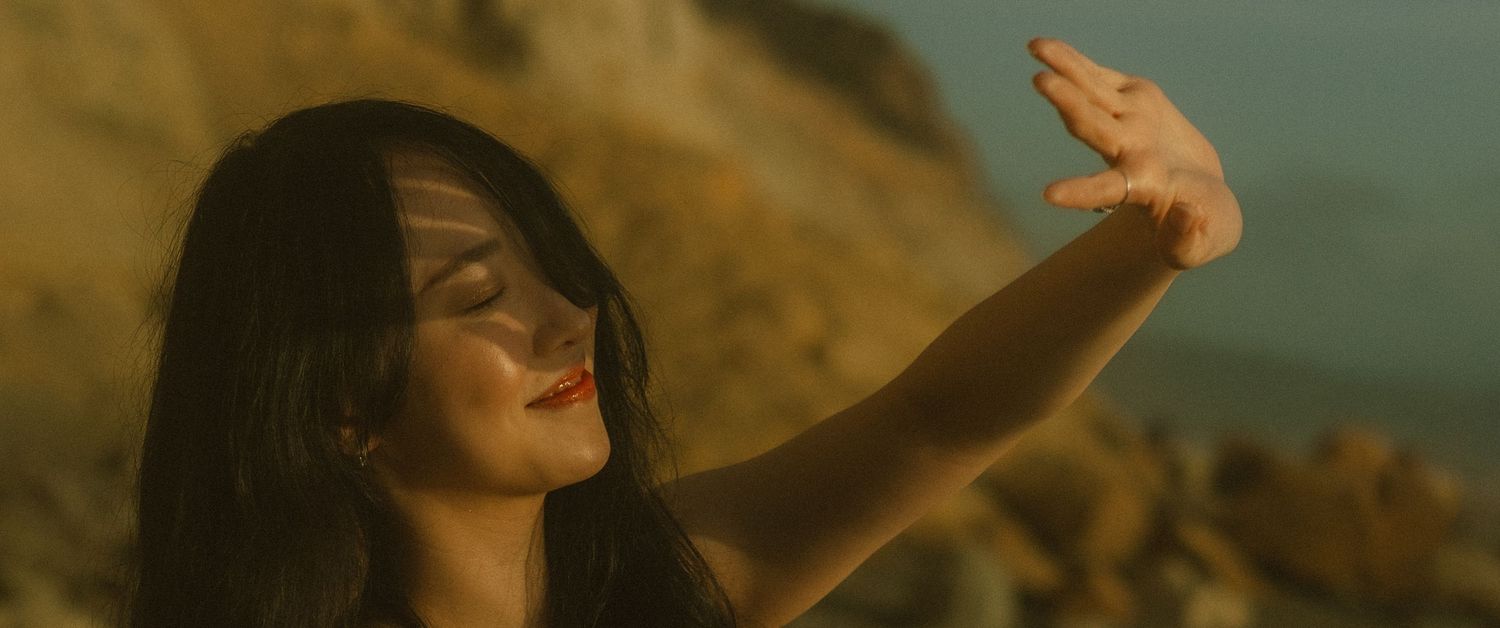 Person with long dark hair reaching towards sunset sky against coastal cliffs.