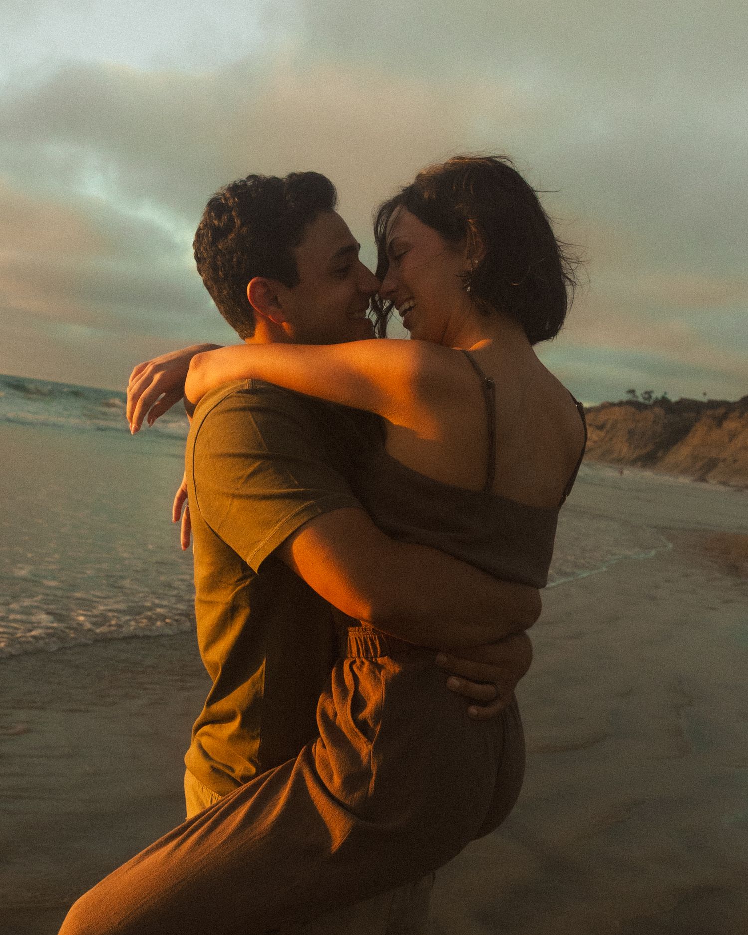 Couple embraces on beach at sunset with golden light illuminating their silhouettes against ocean backdrop.