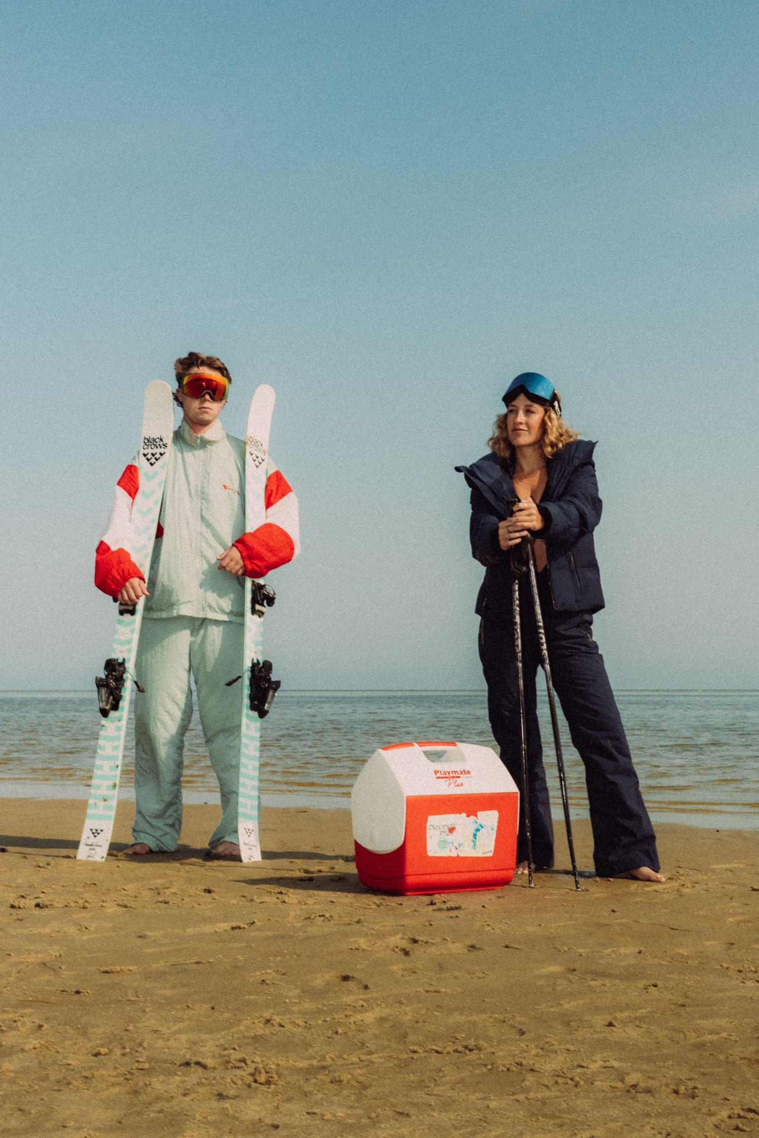 Two people in ski gear pose on a sandy beach with skis, poles, and a cooler under a clear blue sky.
